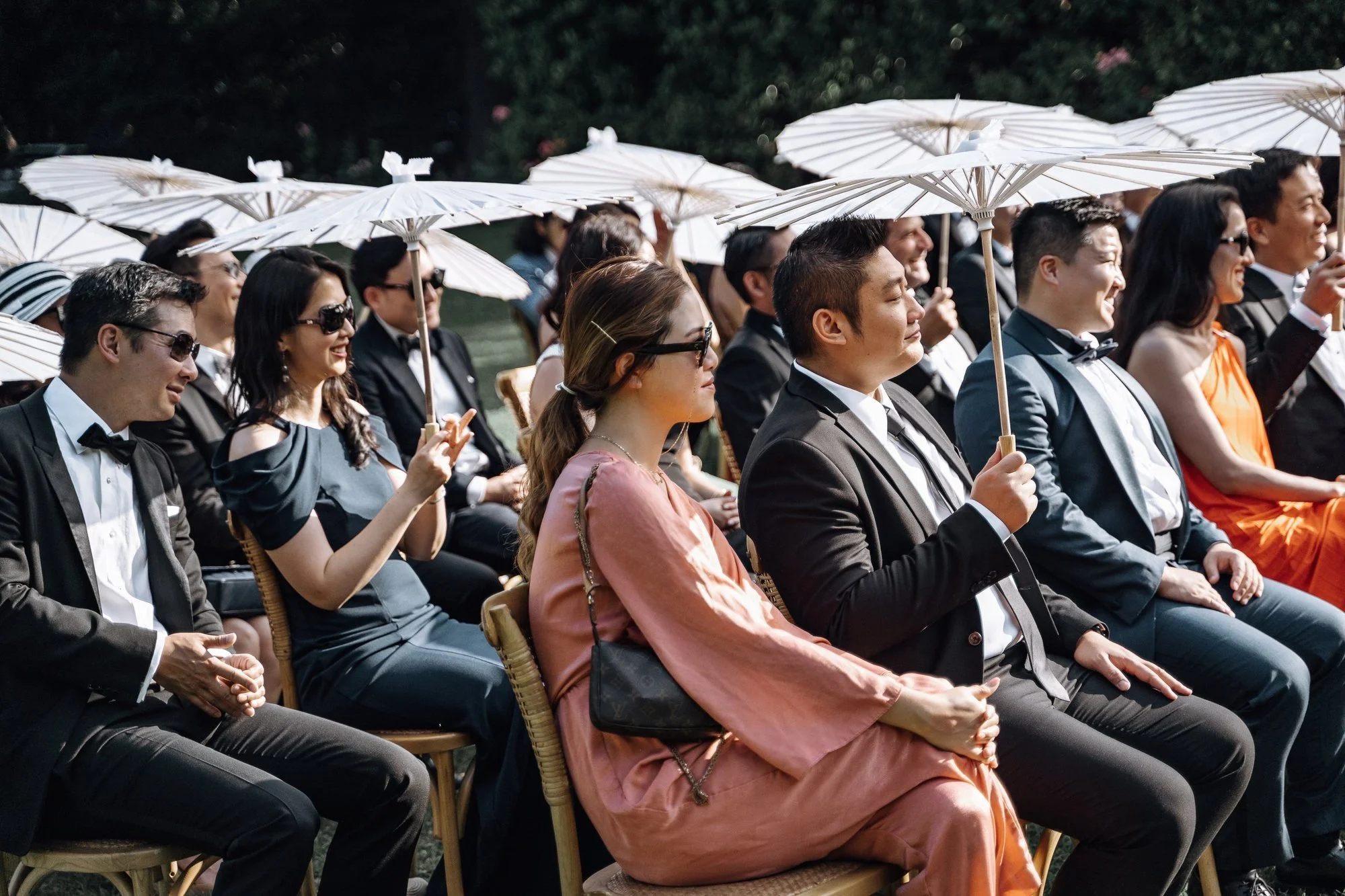 People sitting and smiling at an outdoor event, holding white parasols.