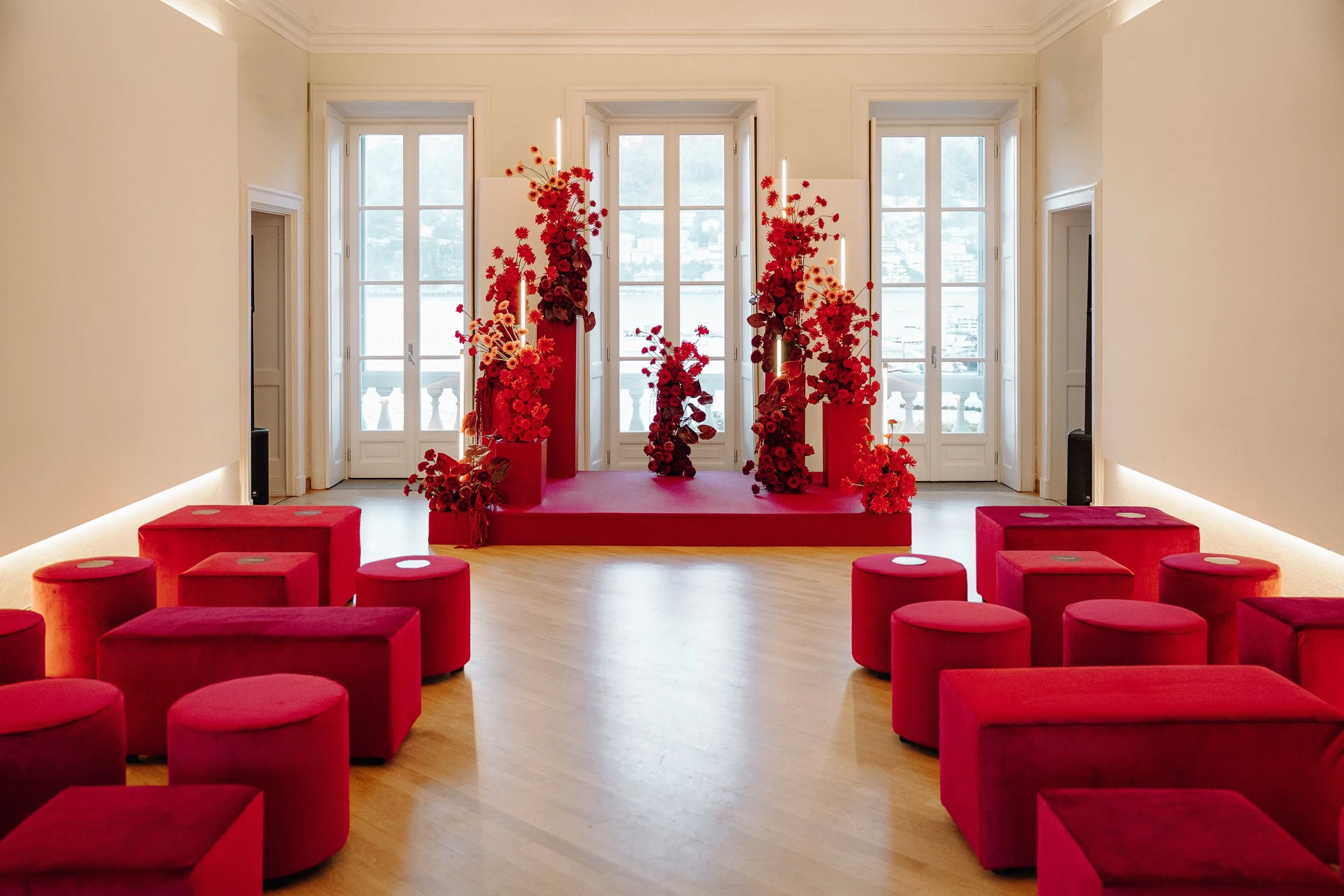 A room with large windows, red floral arrangements on pedestals, red cushioned seats arranged in a semi-circle, and a red carpeted stage area at the front.