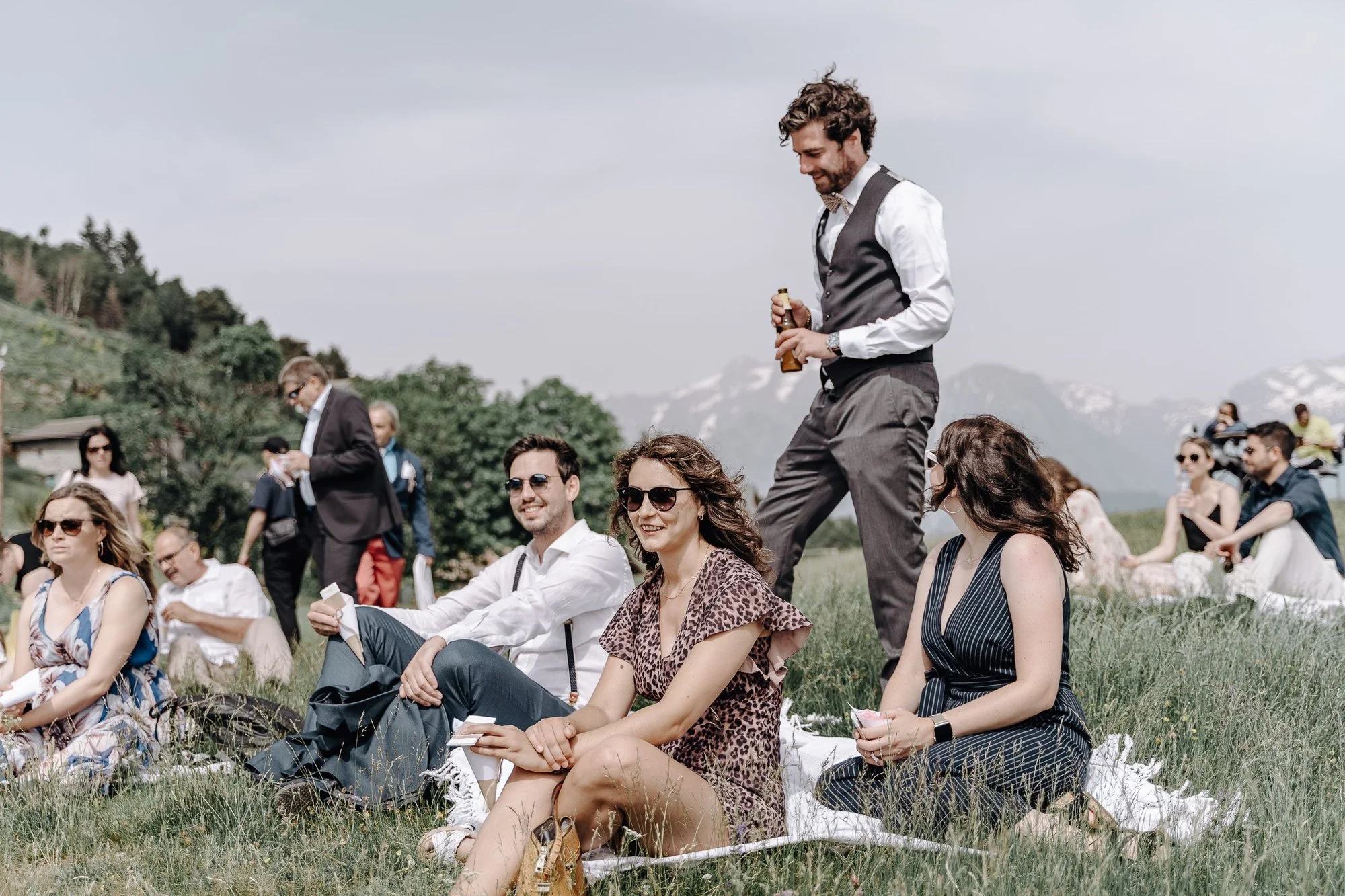 Group of people sitting on grass outdoors during a wedding or event with mountains in the background, some dressed casually and others more formally.