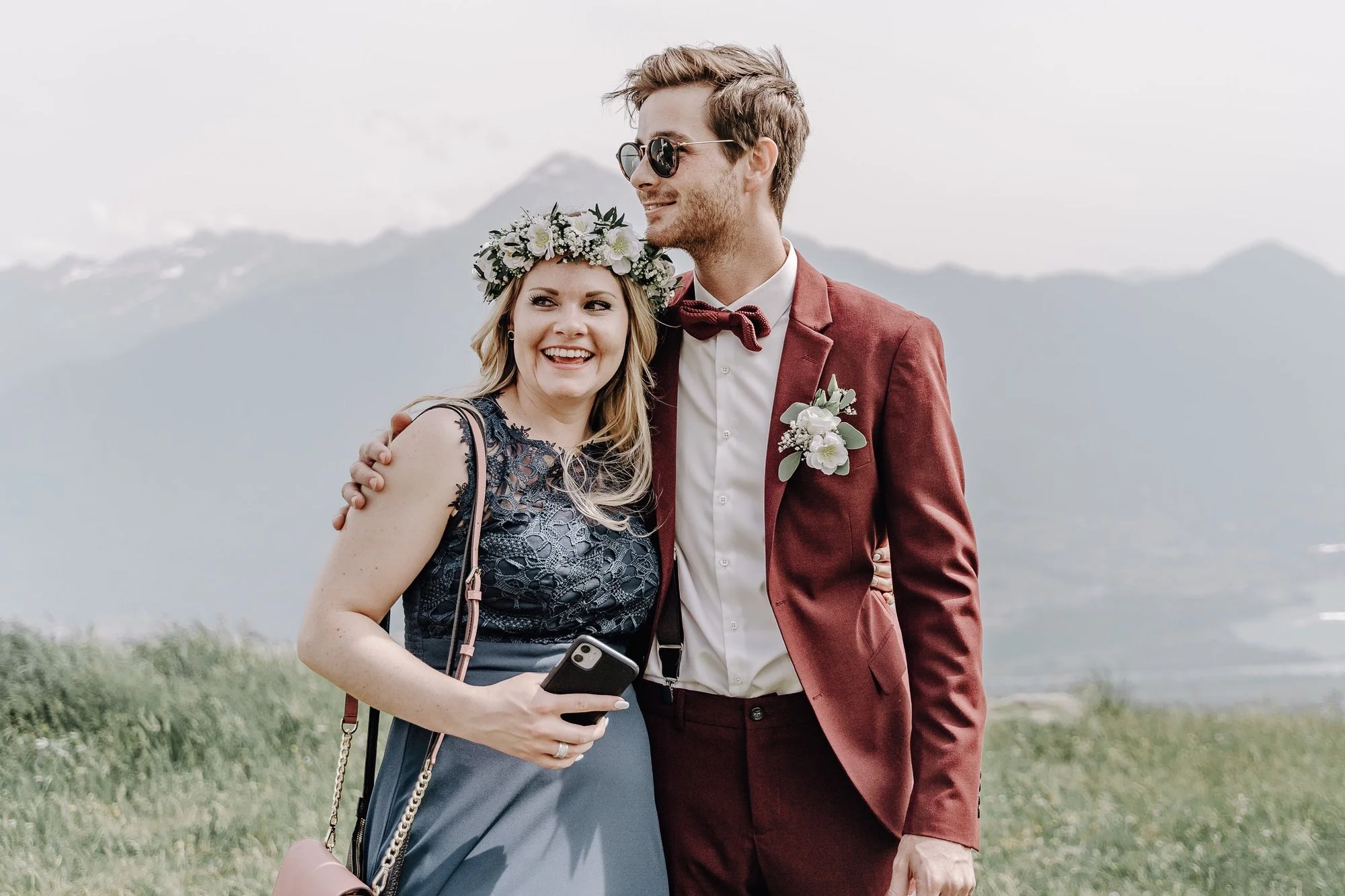 A smiling woman with a floral headband holding a smartphone, and a man in sunglasses and a burgundy suit with a boutonniere, standing outdoors with mountains in the background.