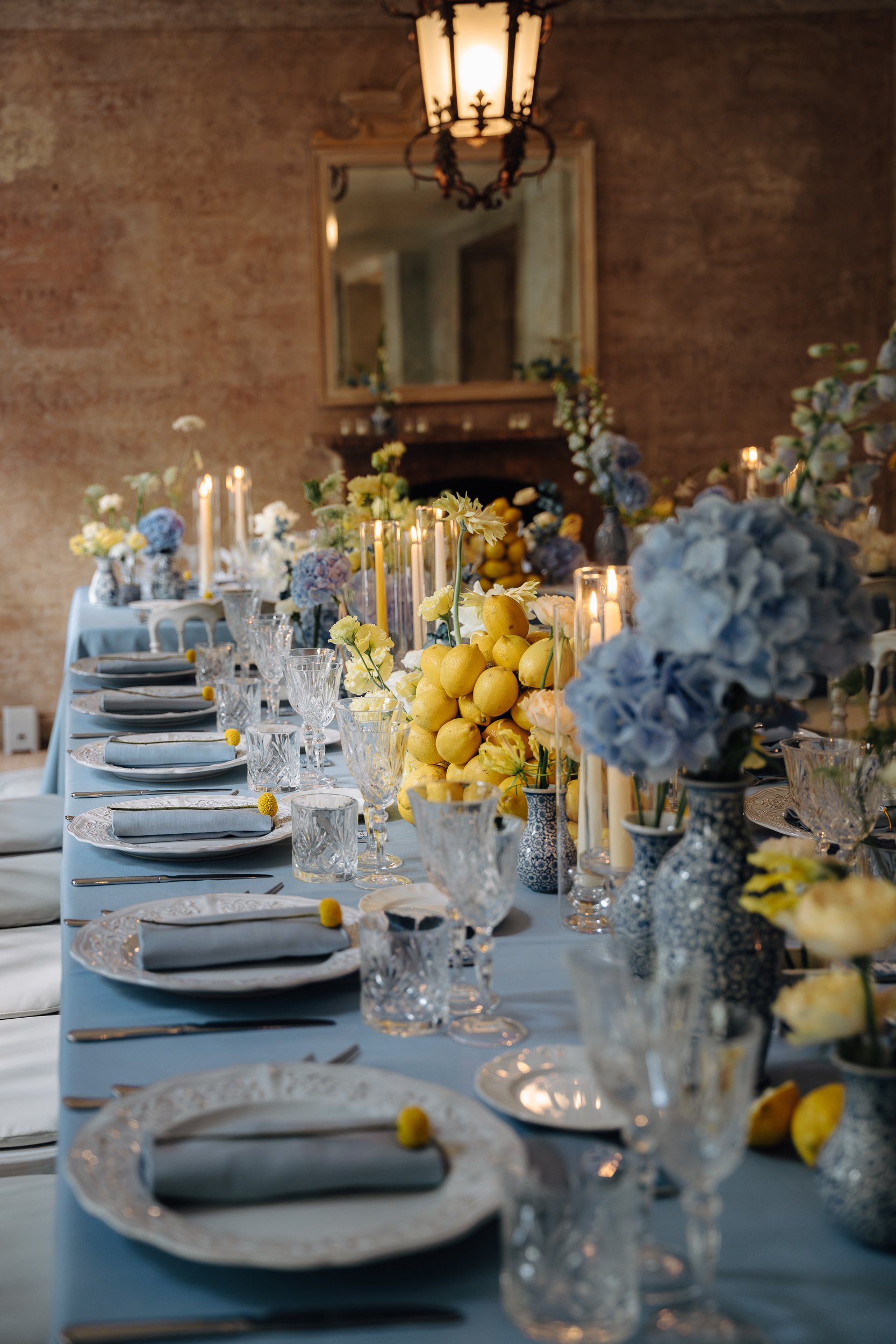 A long banquet table decorated with white and yellow flowers, lemons, candles, and fine glassware, set for a formal event in a room with exposed brick walls and a large mirror.