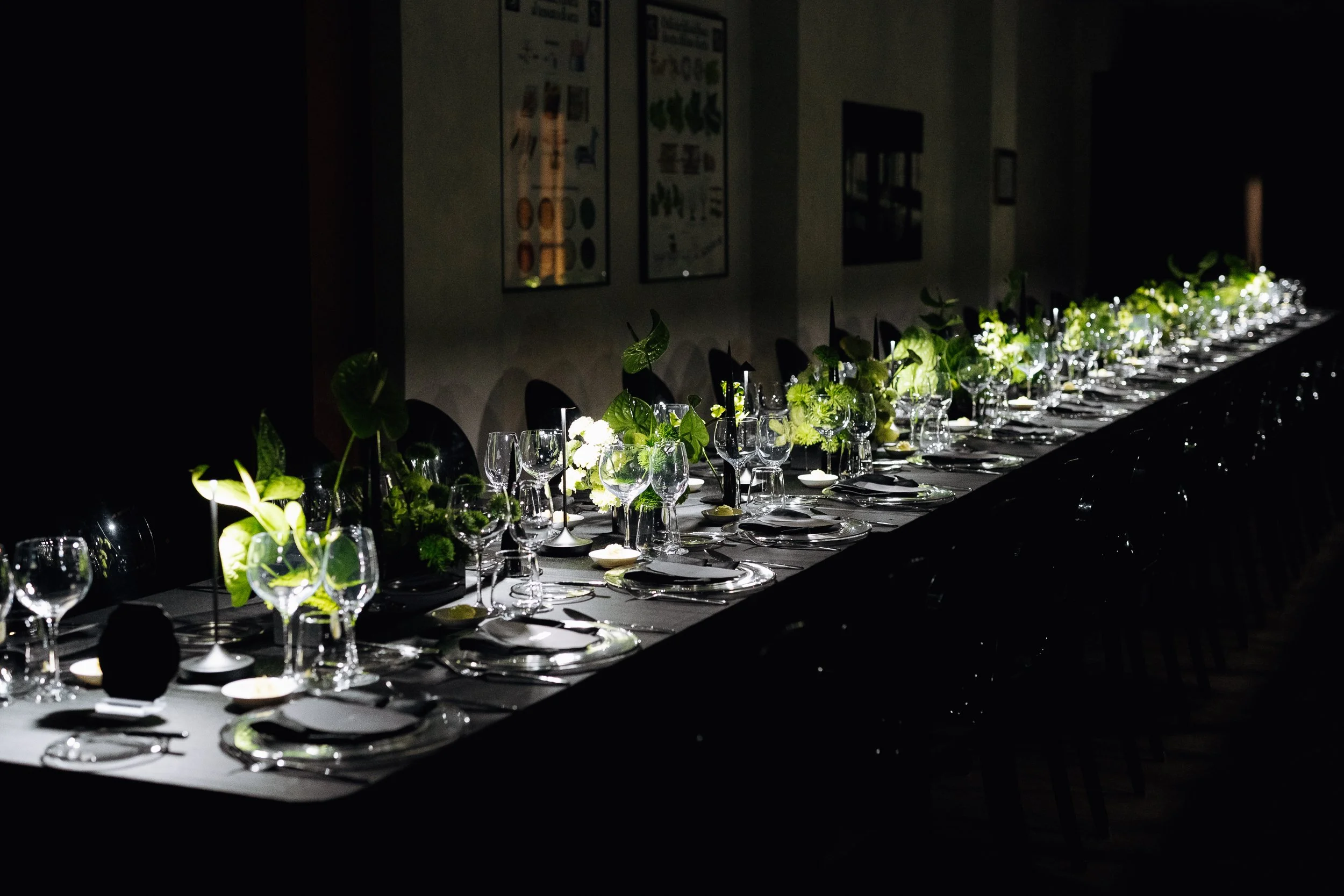 A long dining table set for an event with glassware, black napkins, and greenery as centerpieces. The room is dimly lit, with framed artwork on the wall in the background.