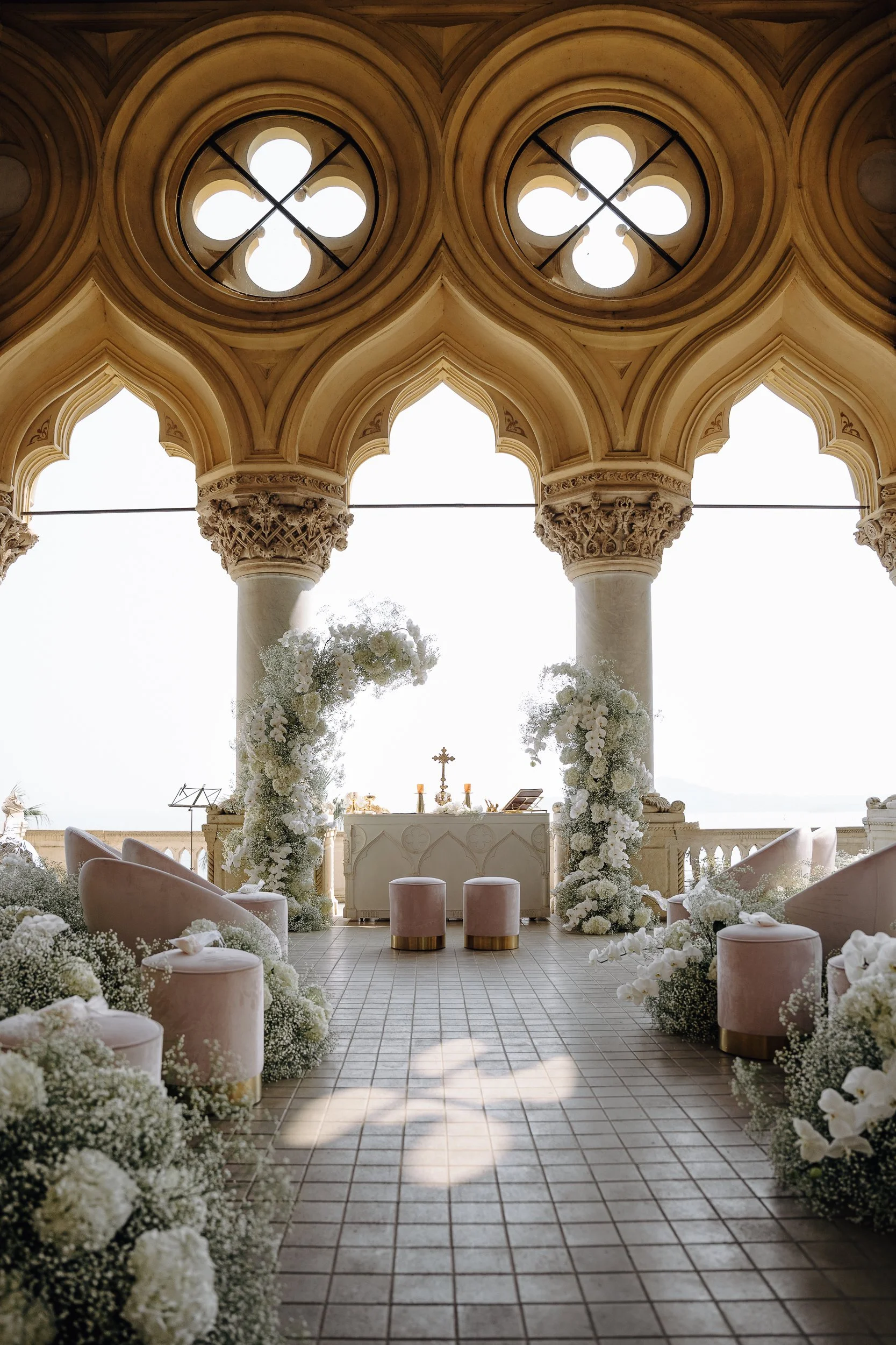 Interior of a wedding altar decorated with white flowers, with a cross and candles behind it, set in a church with arched windows and columns.