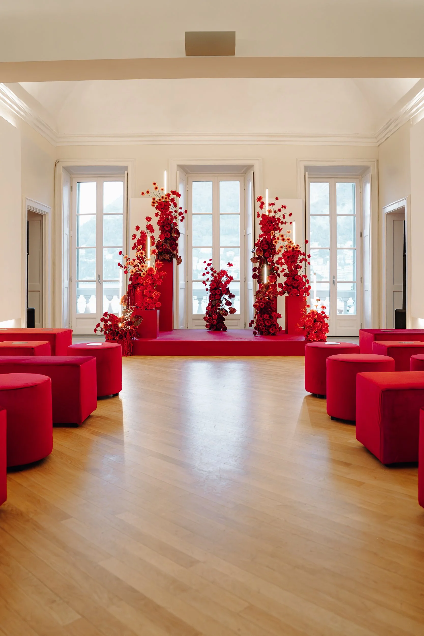 Elegant interior room with large windows, cream-colored walls, and decorative crown molding. There are red floral arrangements on pedestals on a small stage and red upholstered seats arranged around a central open space.