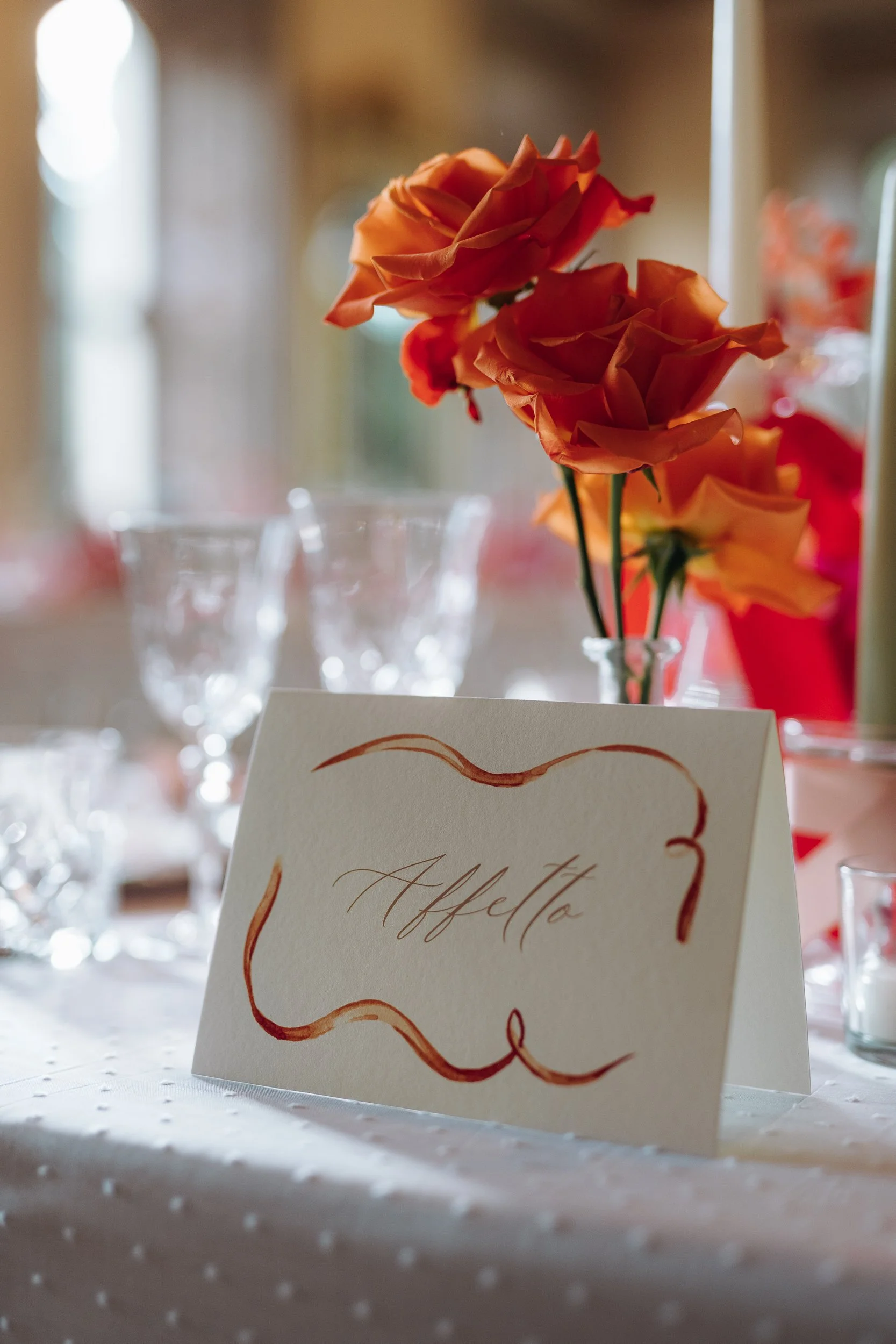 Close-up of a bouquet of orange roses with a place card in the foreground that has the word "Affetto" written on it in cursive, on a table with wine glasses in the background, suggesting a formal event or celebration.