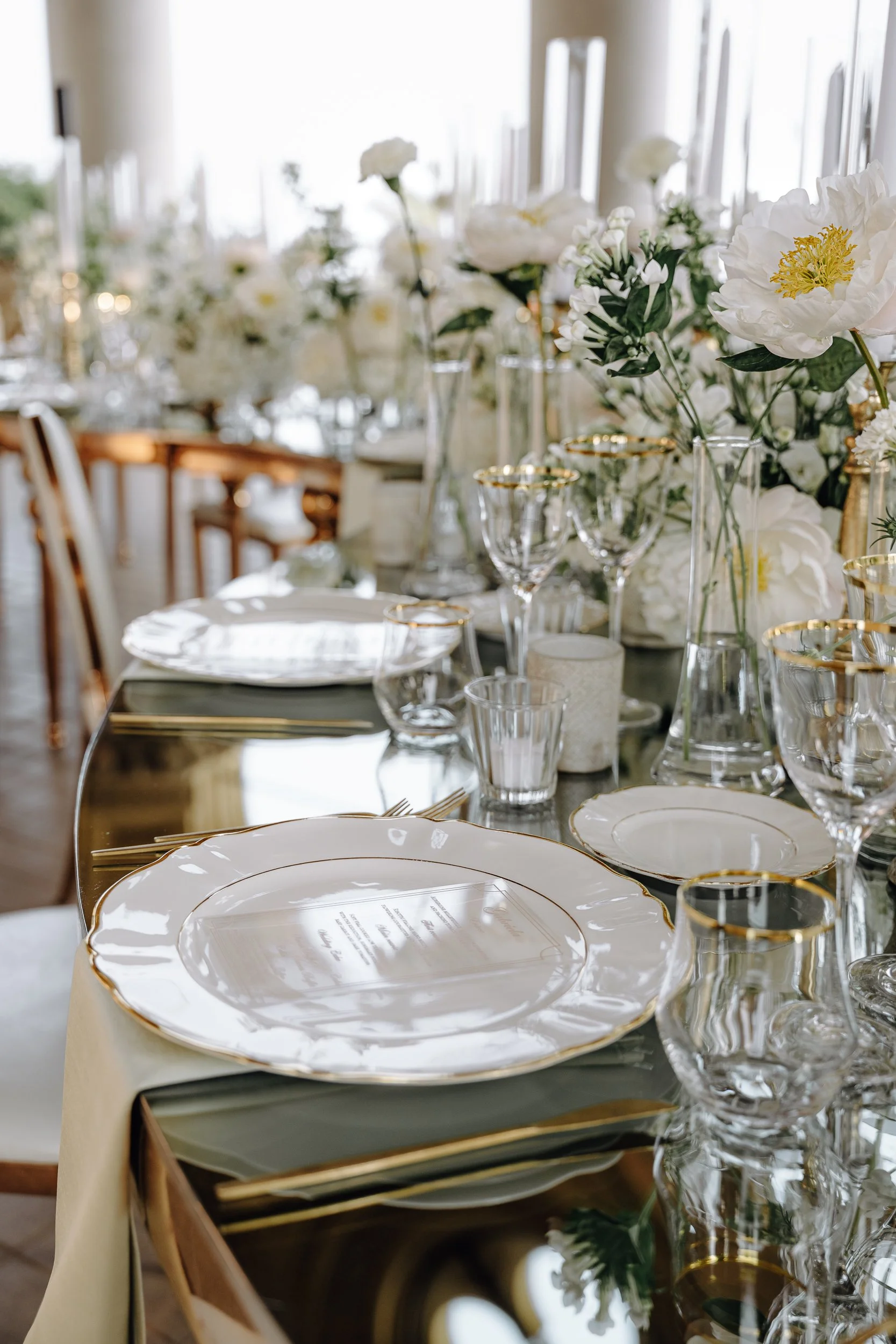 A table set for a formal event with white and gold-rimmed plates, crystal glasses, white flowers, and elegant table decor in a bright room with large windows.