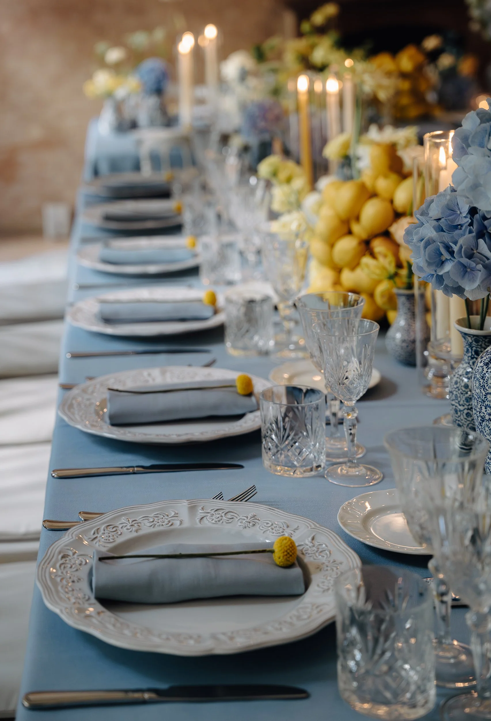A formal dining table set with white plates, crystal glasses, silverware, and rolled napkins with a small yellow flower, decorated with blue and white floral vases, yellow and white flowers, and lit candles, on a light blue tablecloth.