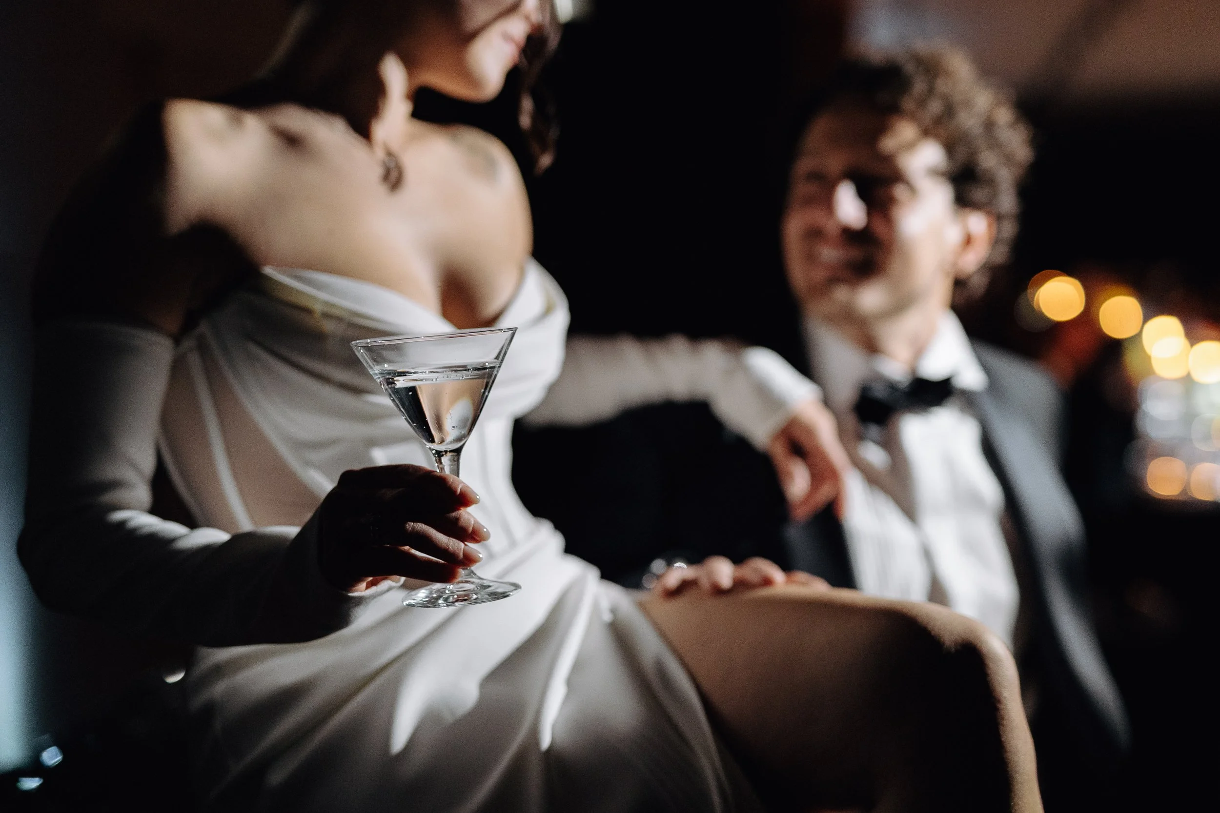 A woman in a white dress holding a cocktail glass, sitting next to a man in a tuxedo, both smiling in a dimly lit setting with bokeh lights in the background.