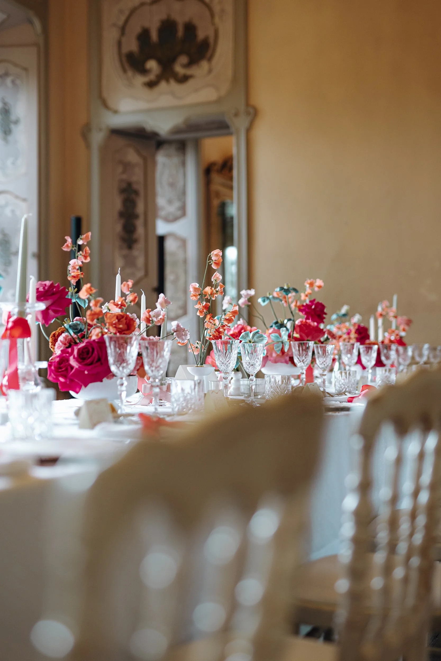 A table decorated with colorful flowers and white candles in a bright room with ornate walls and a mirror.
