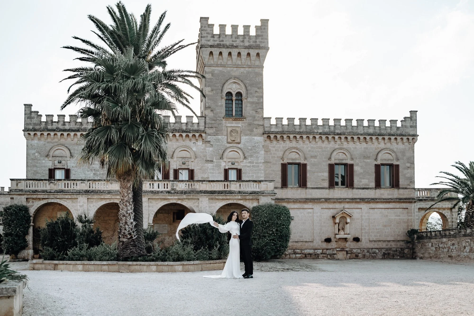 A couple dressed in formal wedding attire standing in front of a historic stone building with a tower, palm trees, and decorative arches. The bride is holding an umbrella.