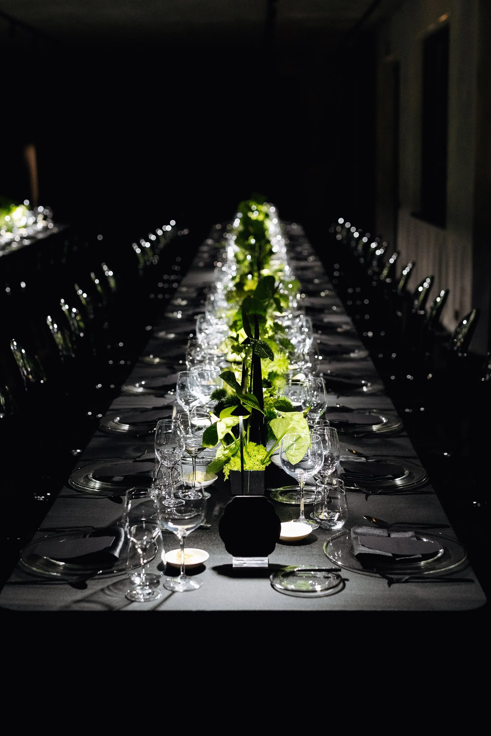 A long dining table set for a formal event with black tablecloths, green floral centerpieces, and glasses arranged with napkins and plates, in a dimly lit room.