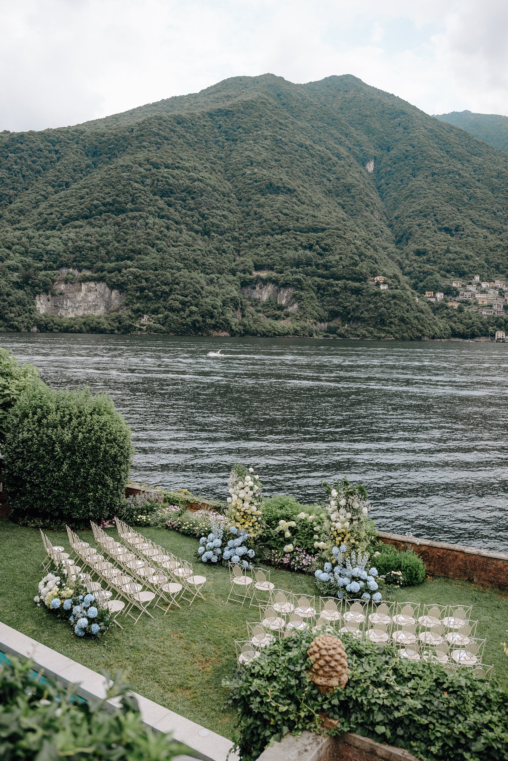 Outdoor wedding setup with white chairs arranged in rows facing a floral altar by a lake, with mountains in the background. Floral arrangements of blue and white flowers decorate the area.
