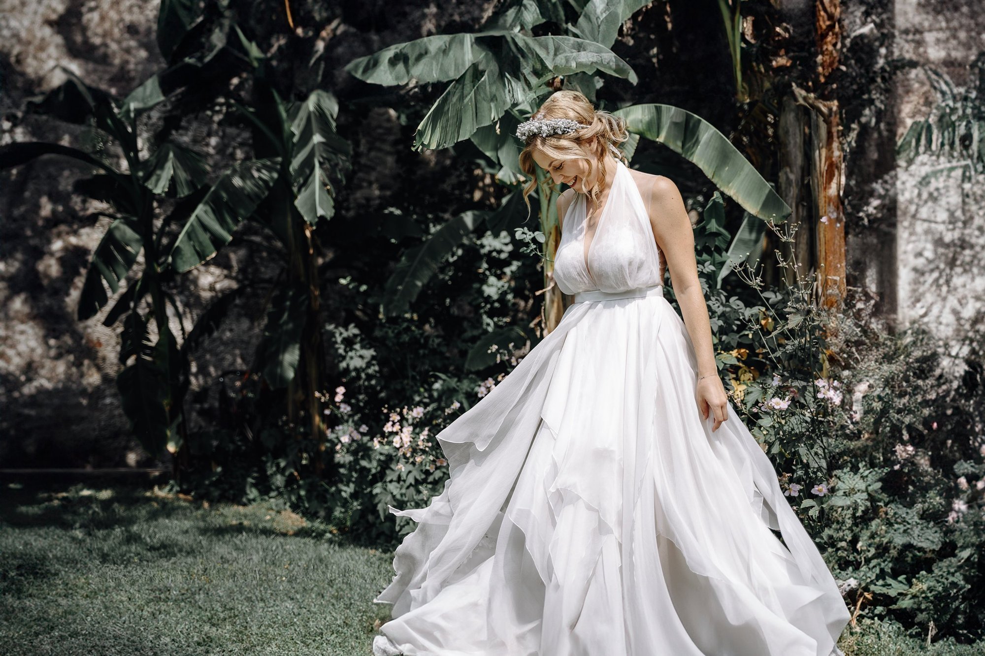 A woman in a white wedding dress with a floral crown is standing outdoors among lush green foliage and flowers, smiling and looking down.
