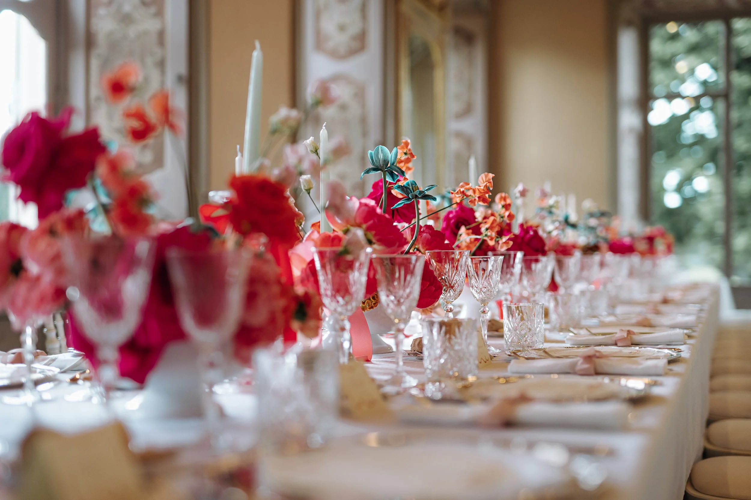 A close-up of a table decorated with pink, red, and white flowers in glass holders, with tall white candles among the flowers, in a room with large windows and ornate decor.