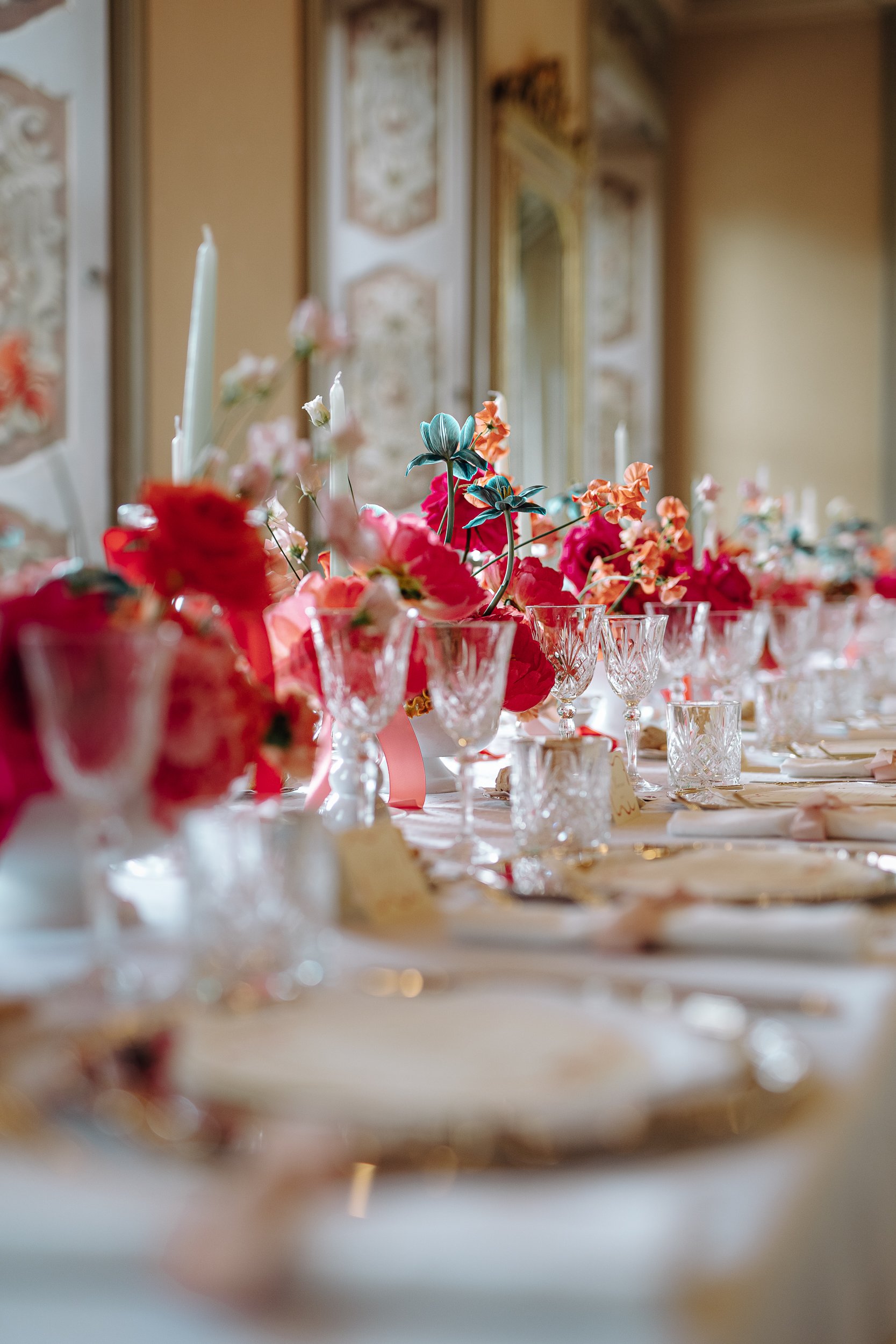 A close-up of a decorated table with glassware and pink and coral flowers, candles, and ribbons, set for a celebration or event in a decorated room.