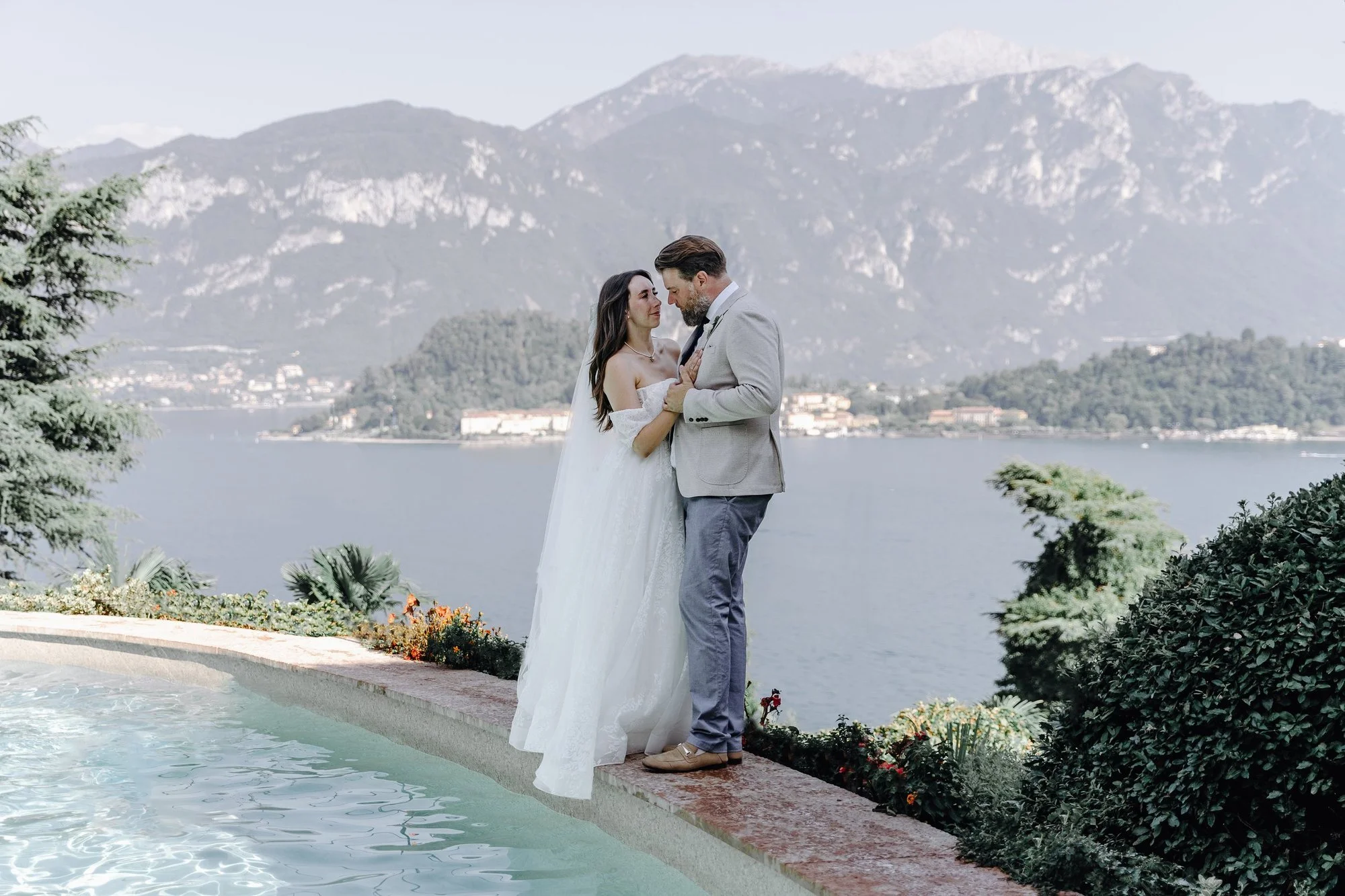 A bride and groom standing by a pool with a mountainous landscape and lake in the background, embracing each other during their wedding in an outdoor setting.