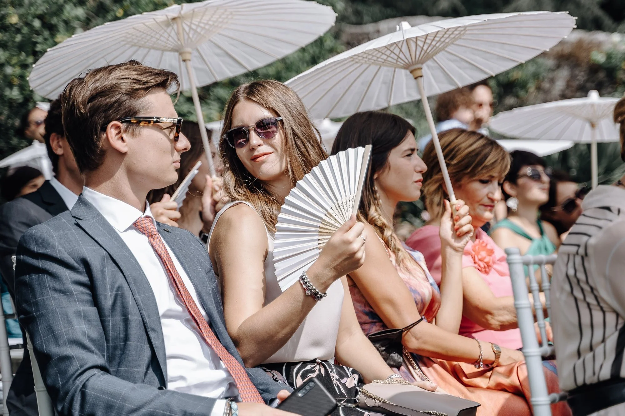 People sitting in chairs outdoors during a daytime event, holding parasols to shield themselves from the sun. A man in a suit and women with sunglasses and fans are visible in the foreground.