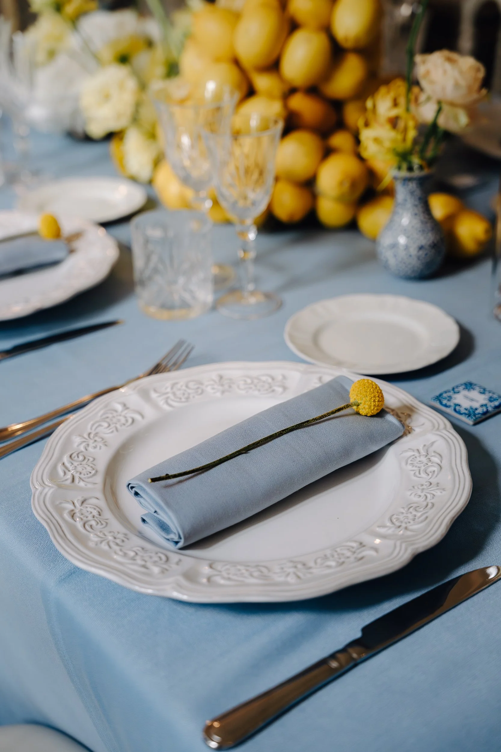 A formal dining table setting with a white ornate plate, a rolled light blue napkin with a yellow billy button flower, gold utensils, a clear glass, and a decorative blue and white vase with flowers in the background.