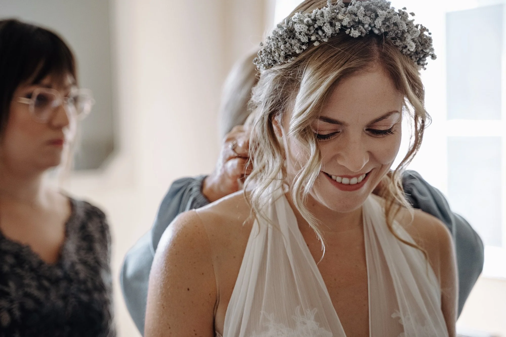 A woman with blonde hair wearing a white dress and a floral crown is smiling as she has her dress adjusted by another woman with dark hair and glasses. The scene is lit by natural light from a window.