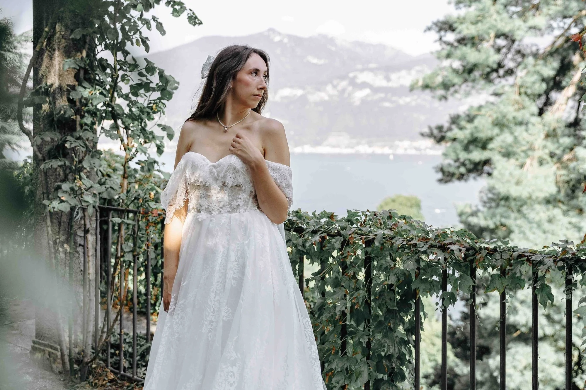A woman in a white off-the-shoulder dress standing by a black wrought iron fence surrounded by green bushes, with a lake and mountains in the background.