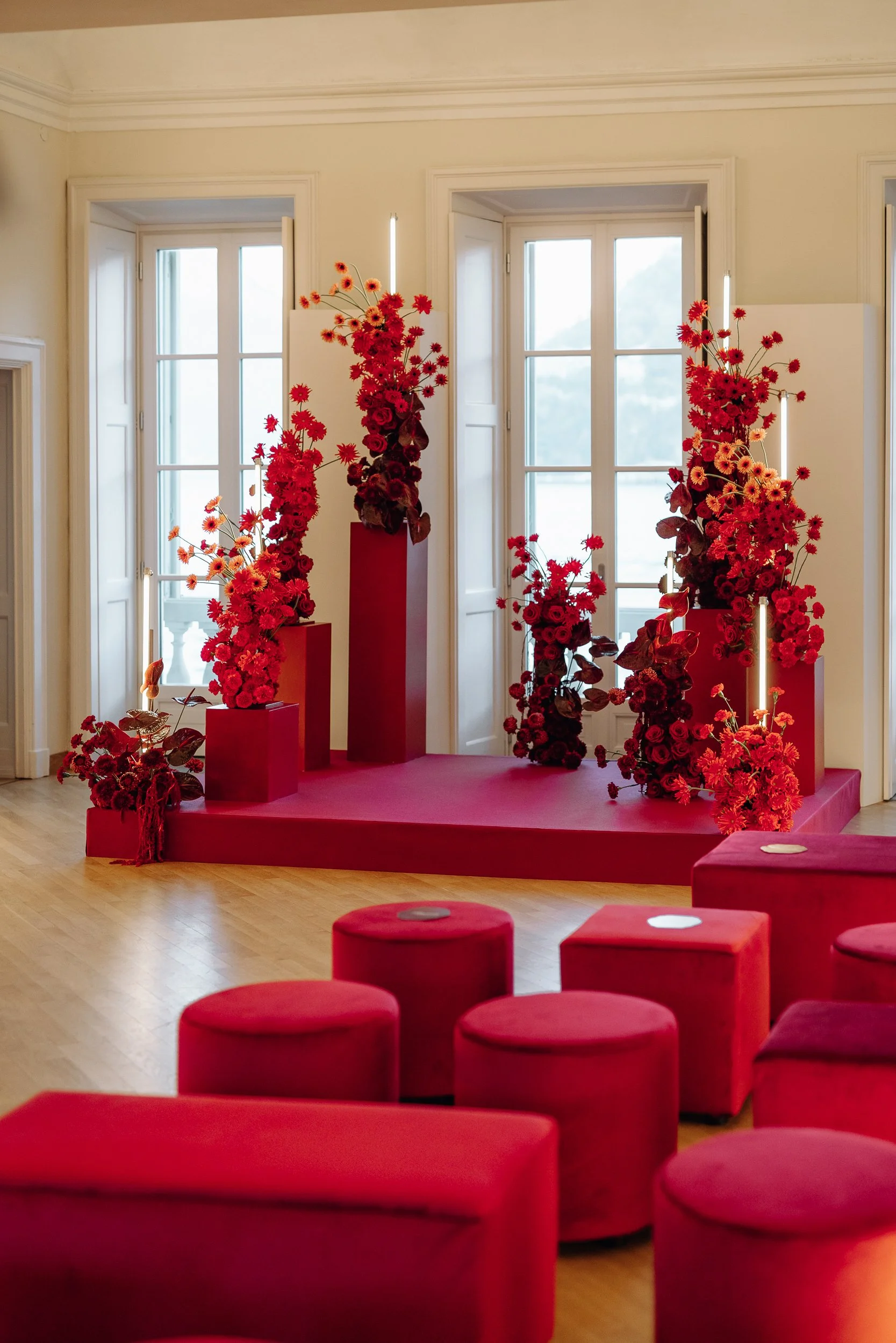 Red floral arrangements on red pedestals in a room with large windows and red cushioned seating.