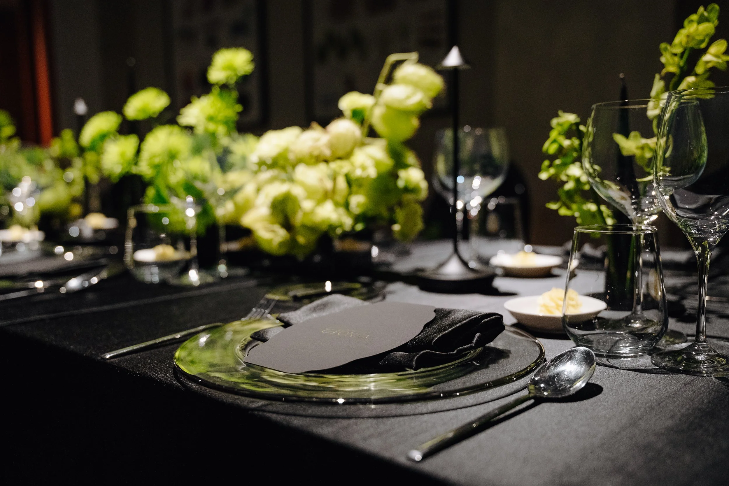 A dining table set for a formal event with floral centerpieces, wine glasses, and plates on a dark tablecloth, in a dimly lit room.