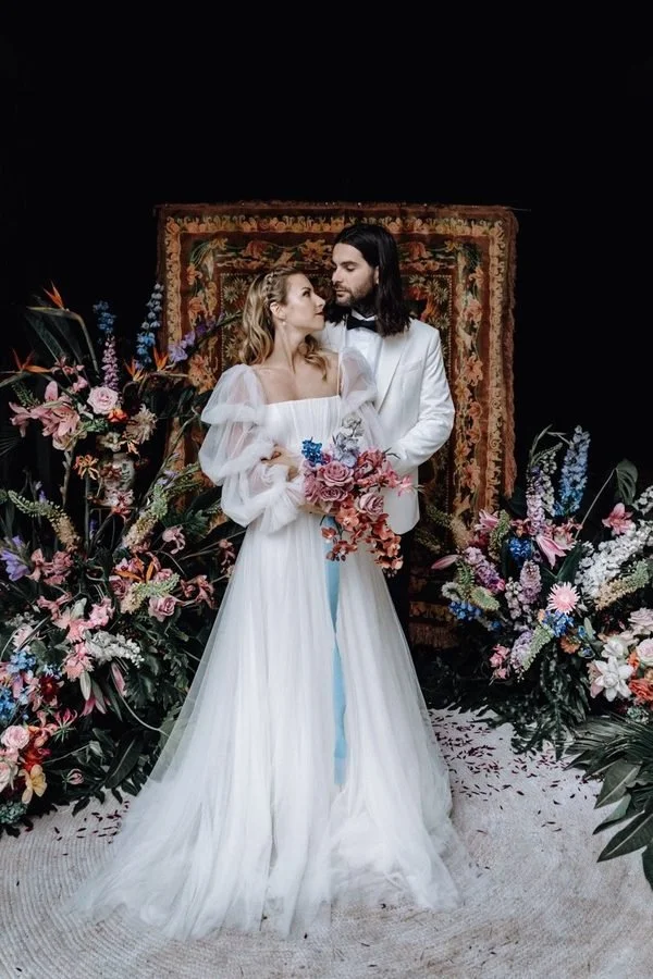 Bride and groom in wedding attire standing amidst colorful flowers, with a decorative rug backdraft, holding a bouquet of flowers, in a wedding setting.