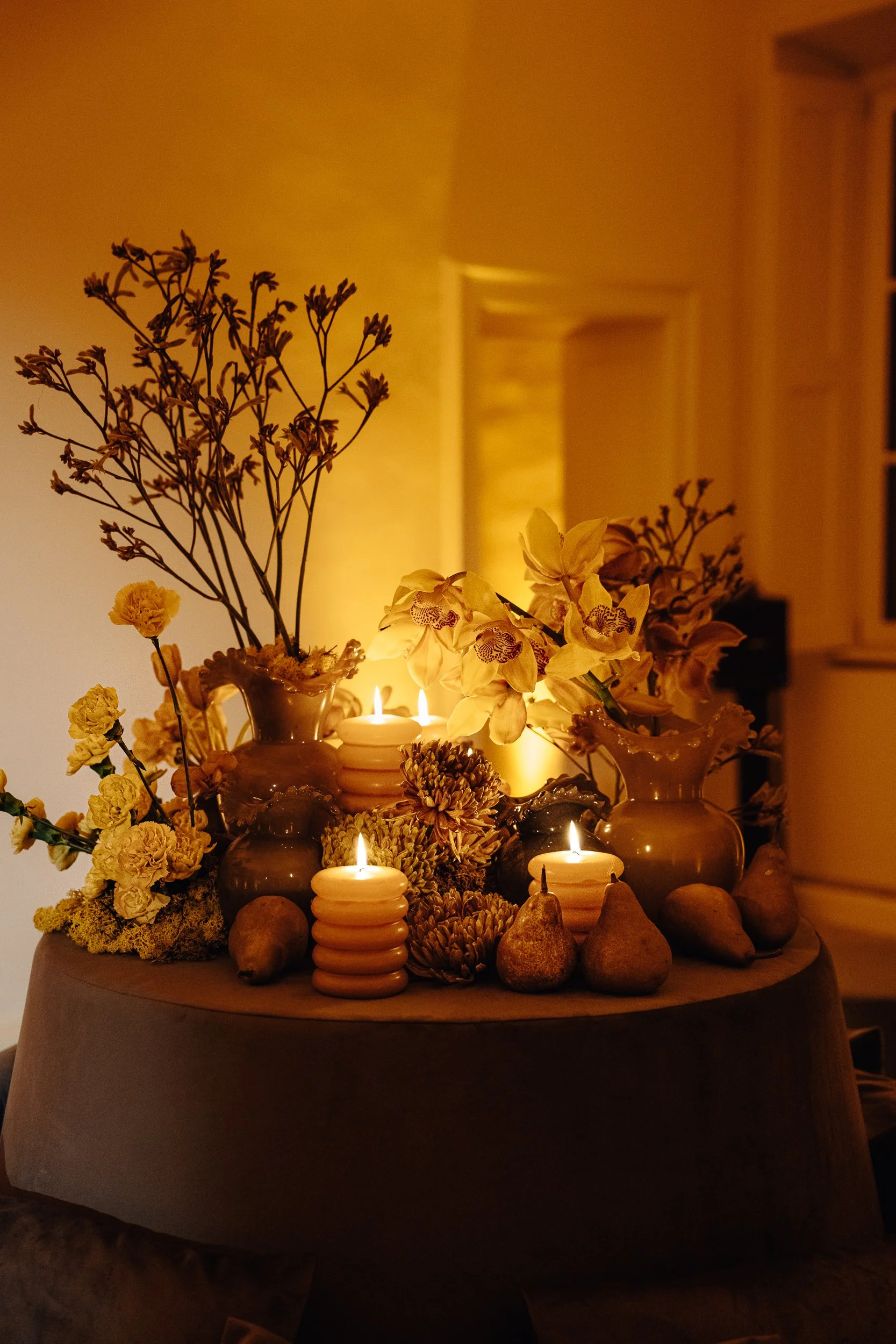 A decorative table with multiple lit candles, surrounded by assorted flowers and fruits, in a warm, softly lit room.