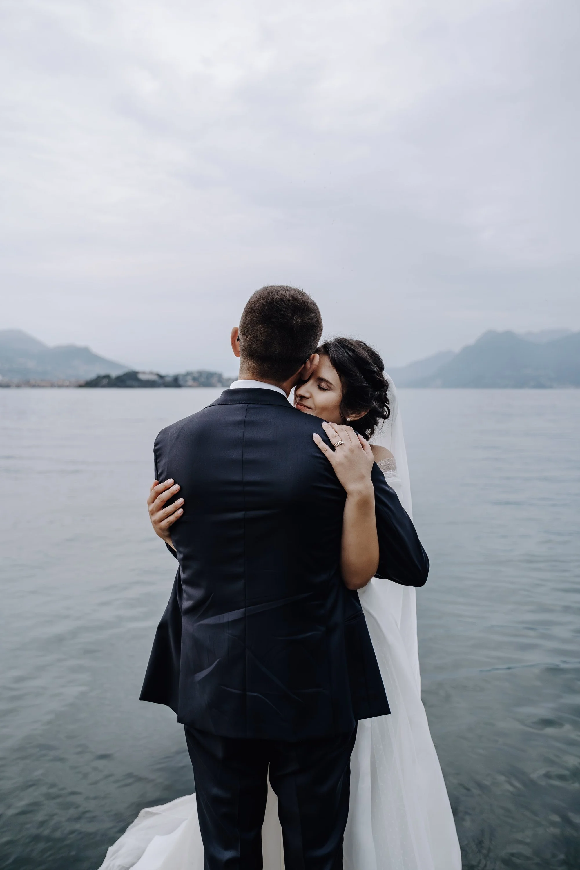 A bride and groom embracing by a body of water with mountains in the background. The groom is in a dark suit, and the bride has dark hair with a veil, and is wearing a white wedding dress.