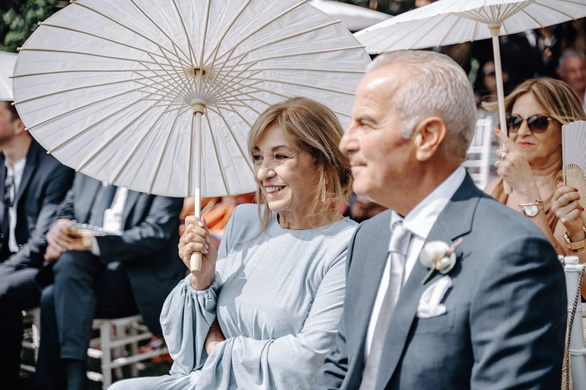 People sitting outdoors, some using white umbrellas for shade. The focus is on a woman with light brown hair, smiling and holding a white parasol, and a man with gray short hair, dressed in a gray suit, sitting next to her with arms crossed.
