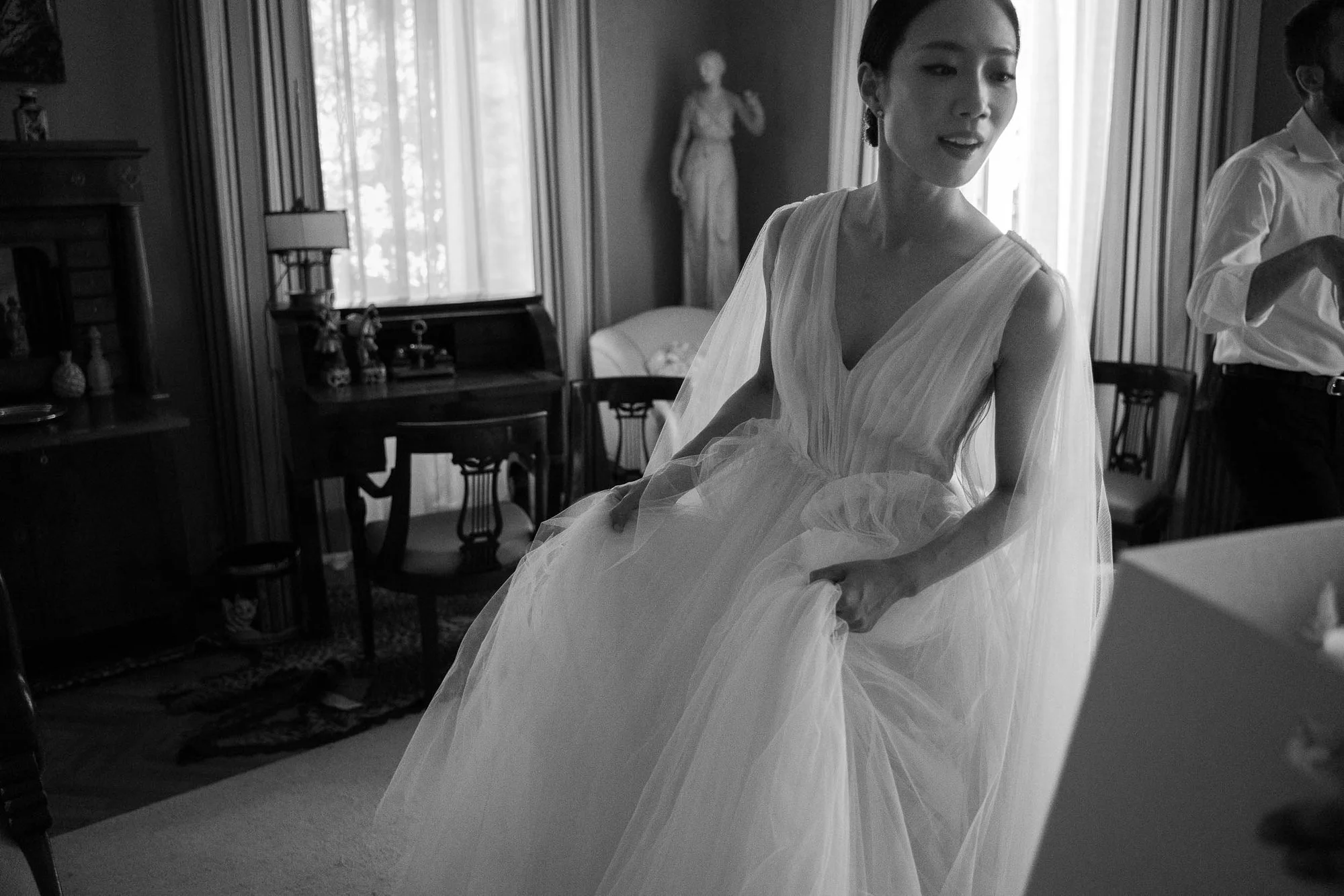 A woman in a flowing wedding dress sitting in a room with large windows and curtains, with a man standing beside her and a woman in the background.