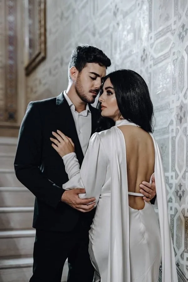 A man and woman standing close together indoors, with the man in a suit and the woman in a backless white dress, both with their eyes closed and emotional expressions.
