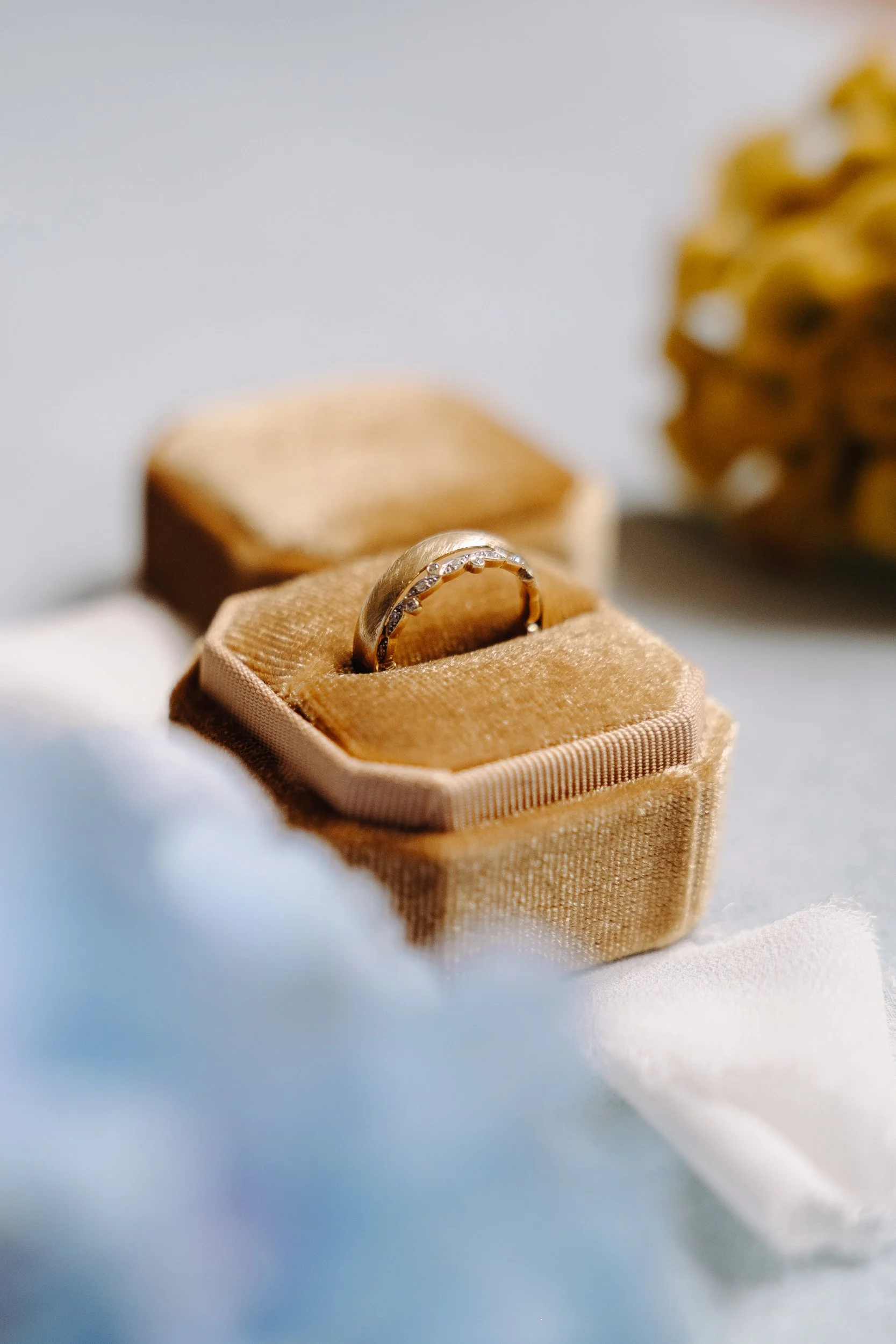 A gold ring with small diamonds in a beige velvet jewelry box, with a blurred background of other jewelry and a yellow flower.