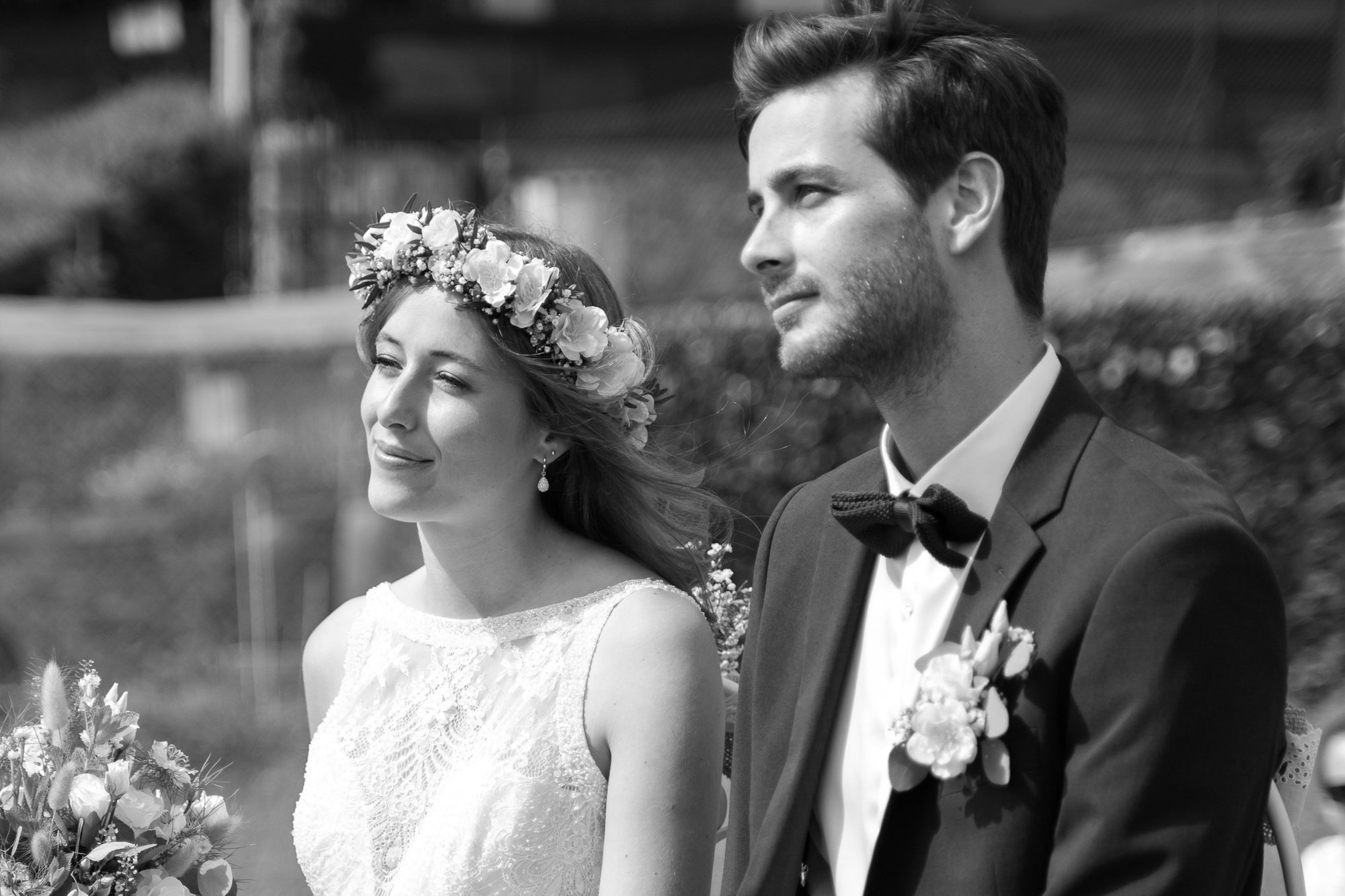 Black and white photograph of a bride and groom at a wedding. The bride, with light skin, is wearing a lace wedding dress and a floral crown. The groom, with light skin, is dressed in a tuxedo with a bow tie and boutonniere. They are standing outdoor