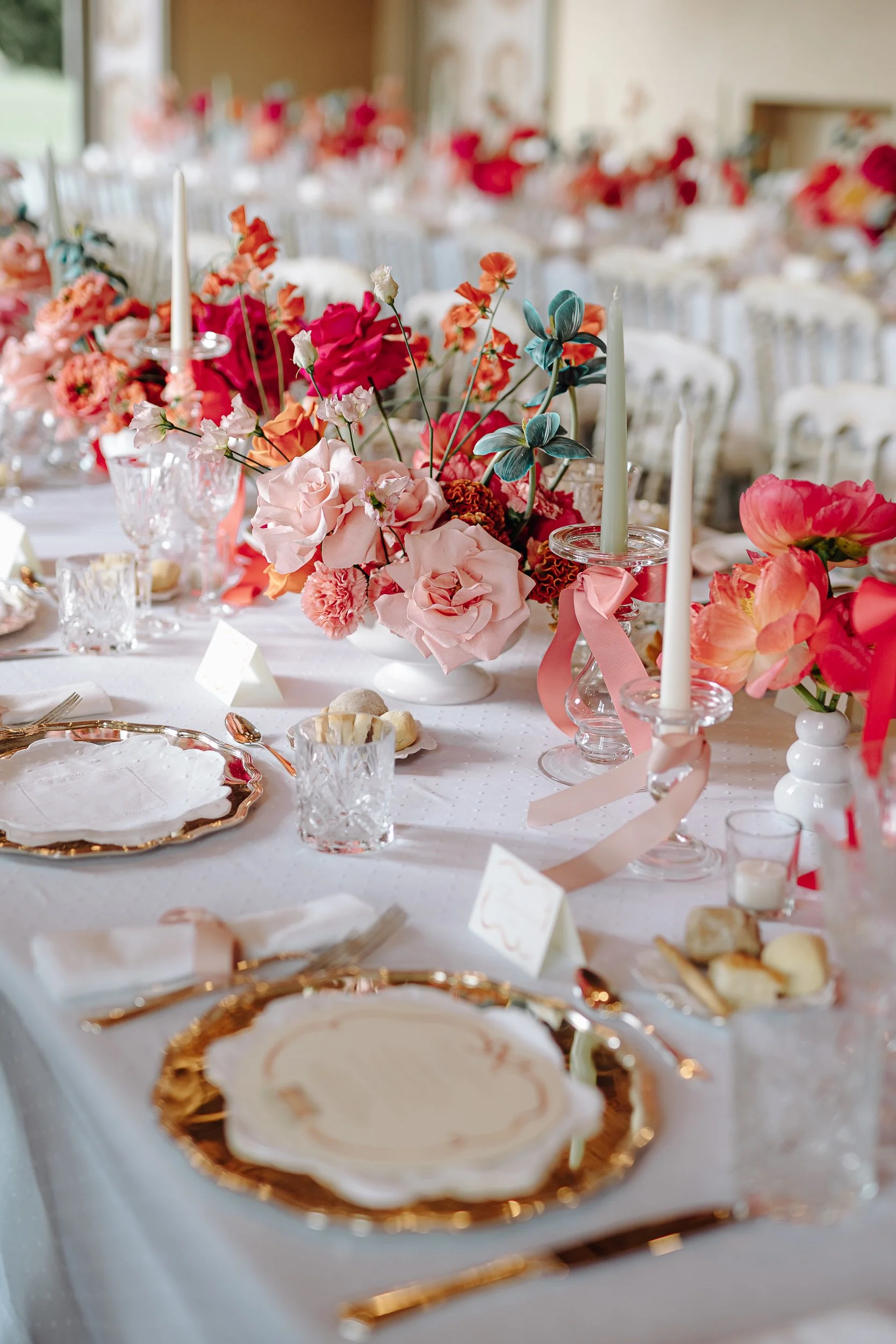 A decorated banquet table with floral centerpieces, tall white candles, ornate gold-rimmed plates, and crystal glassware, set for a formal event or wedding reception.