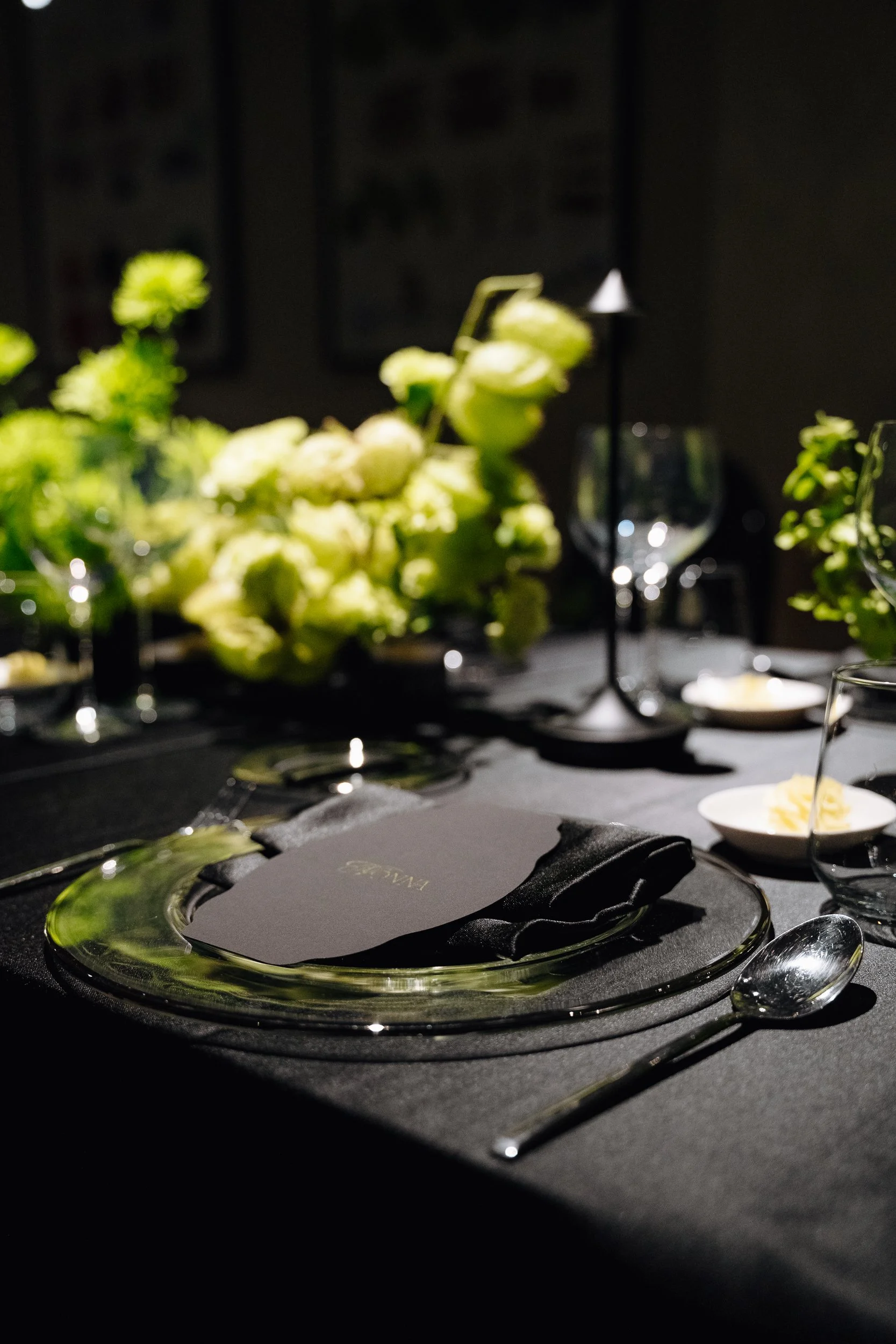 A formal dinner setting with a black tablecloth, elegant glassware, a plate with a black card, and a floral centerpiece of white flowers. In the background, there are two small framed pictures and a black lamp.