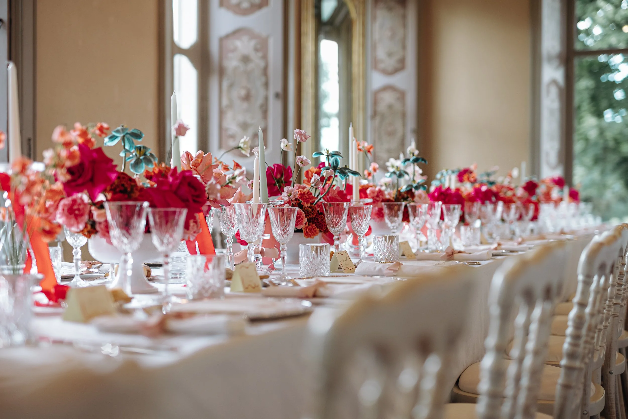 A long banquet table set for a formal event with floral centerpieces, tall candlesticks, and elegant glassware inside a decorated room with large windows.