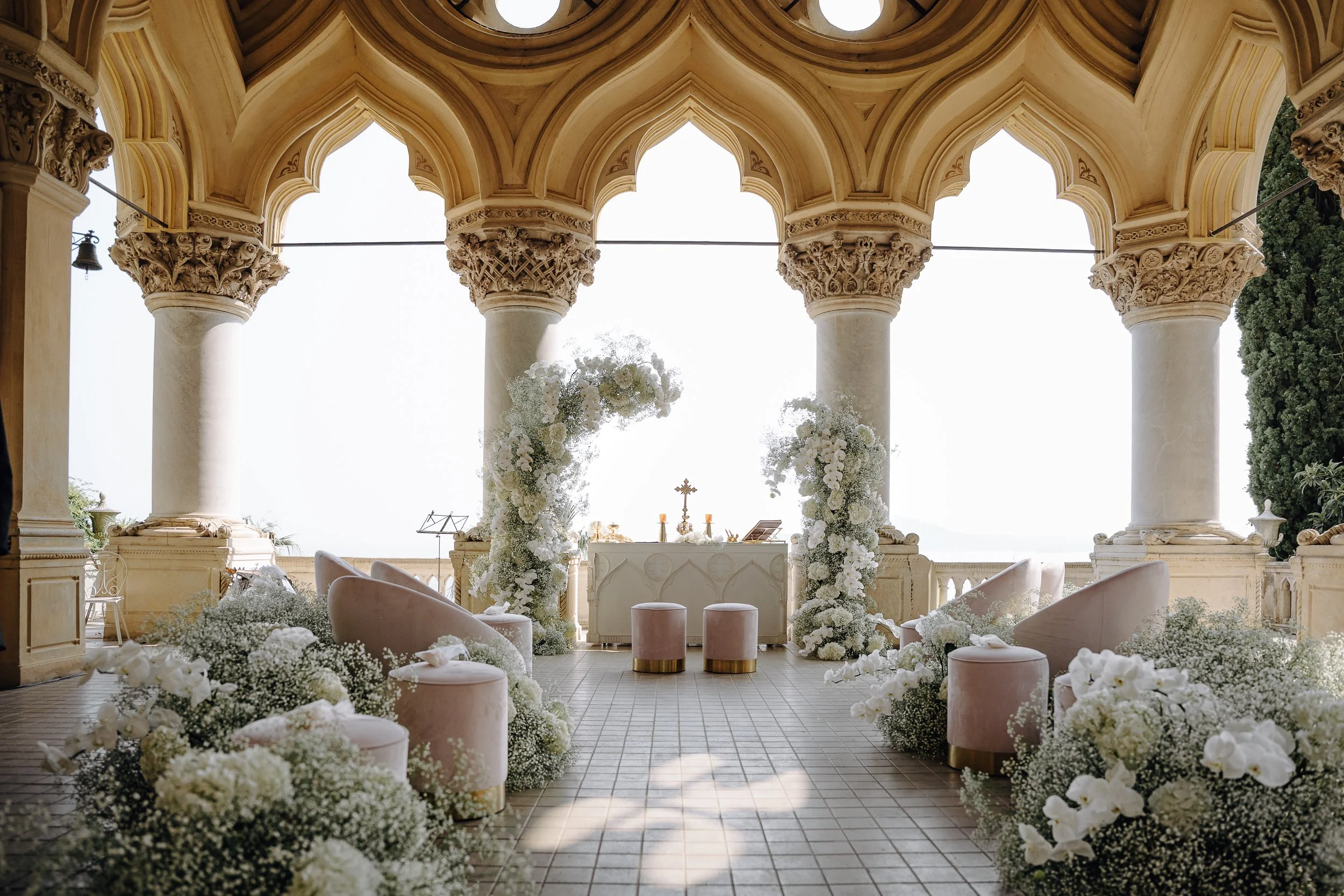 Elegant wedding altar decorated with white flowers and surrounded by columns, with chairs and pink ottomans arranged in front, under a domed ceiling with archways.