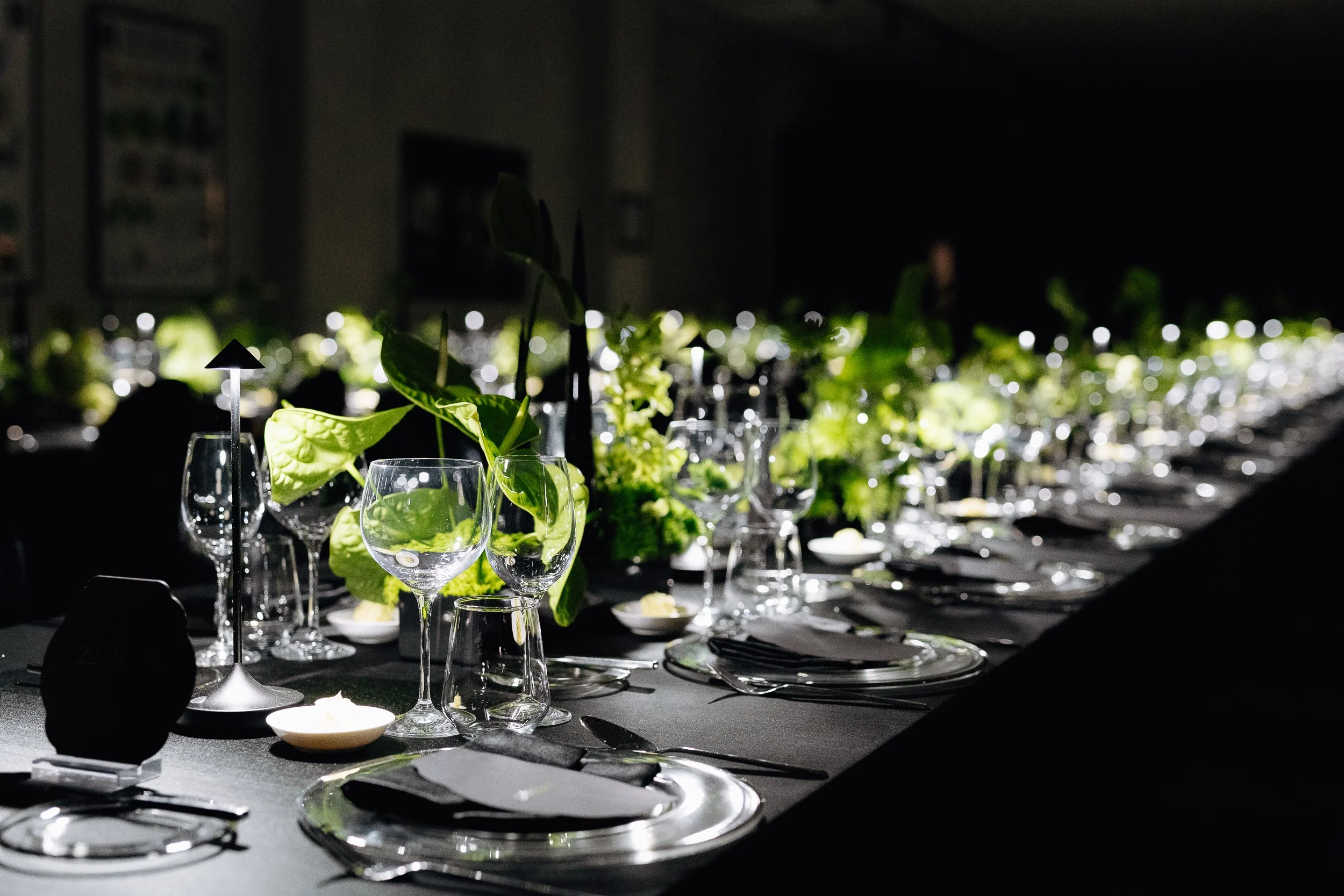 A banquet table set with wine glasses, plates, cutlery, and decorative green foliage with small lights, in a dimly lit room.