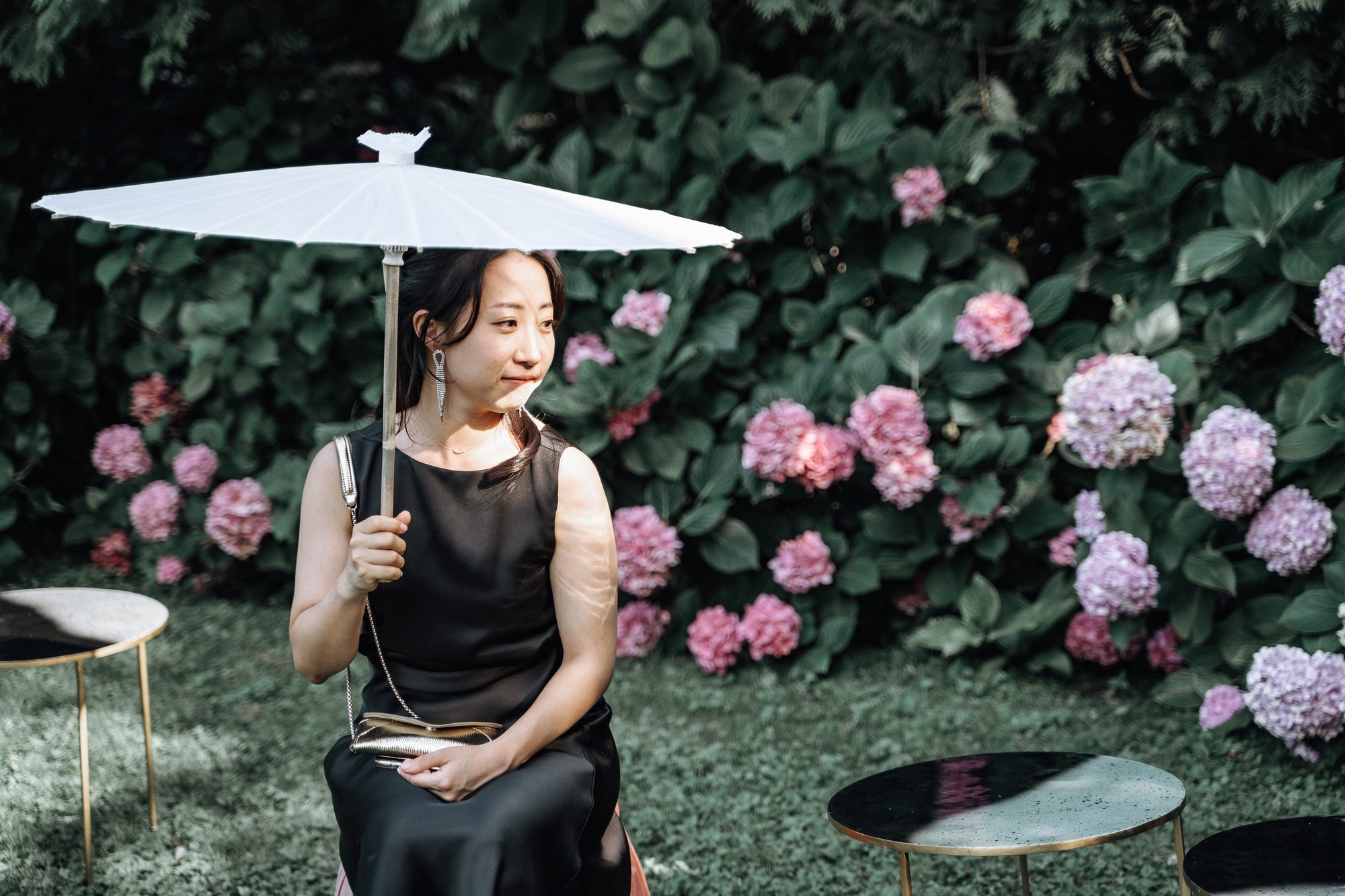 A woman dressed in black sitting outside in front of a hedge with blooming pink and purple hydrangea flowers, holding a white parasol.