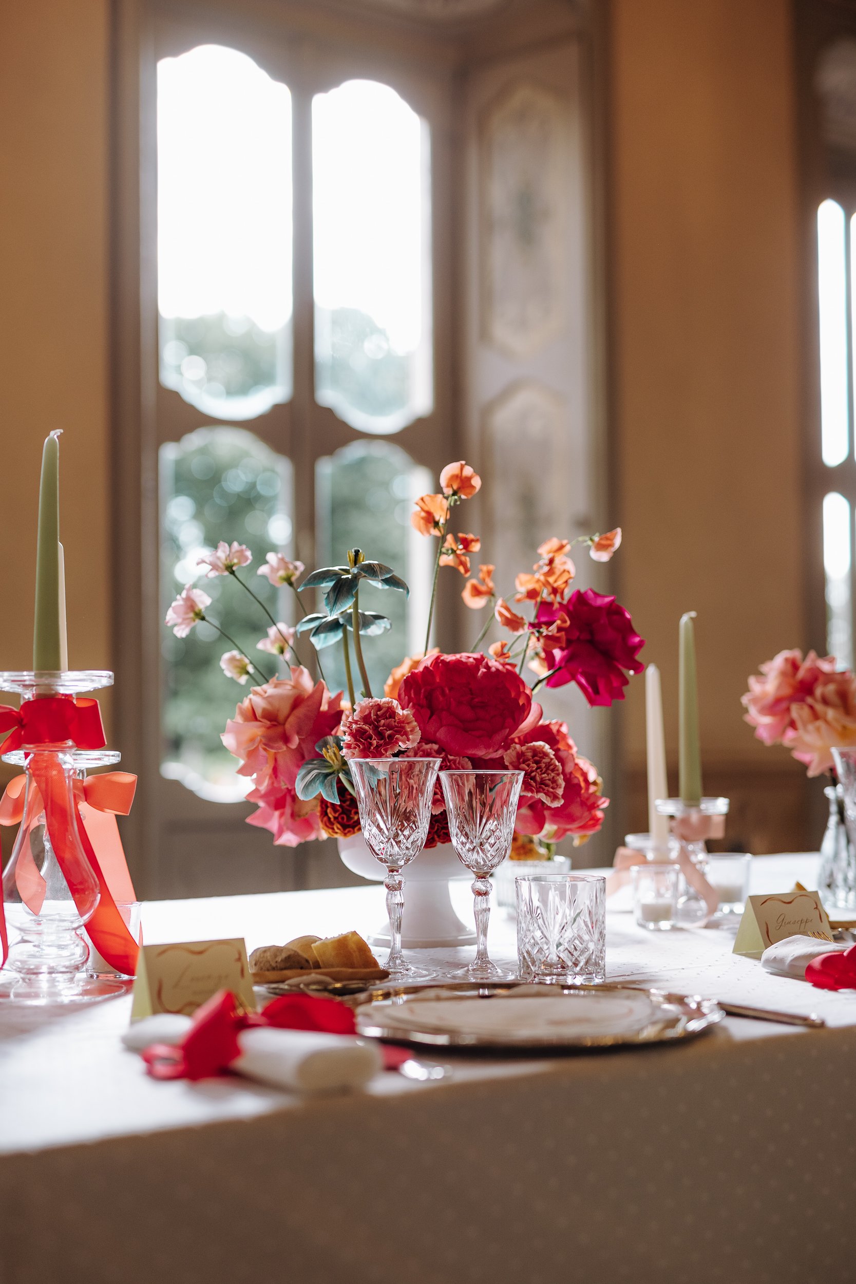 A table decorated with a large bouquet of pink, red, and orange flowers in a white vase, set with glassware, plates, and napkins, in front of a window with ornate wooden frames, suggesting a formal event or celebration.