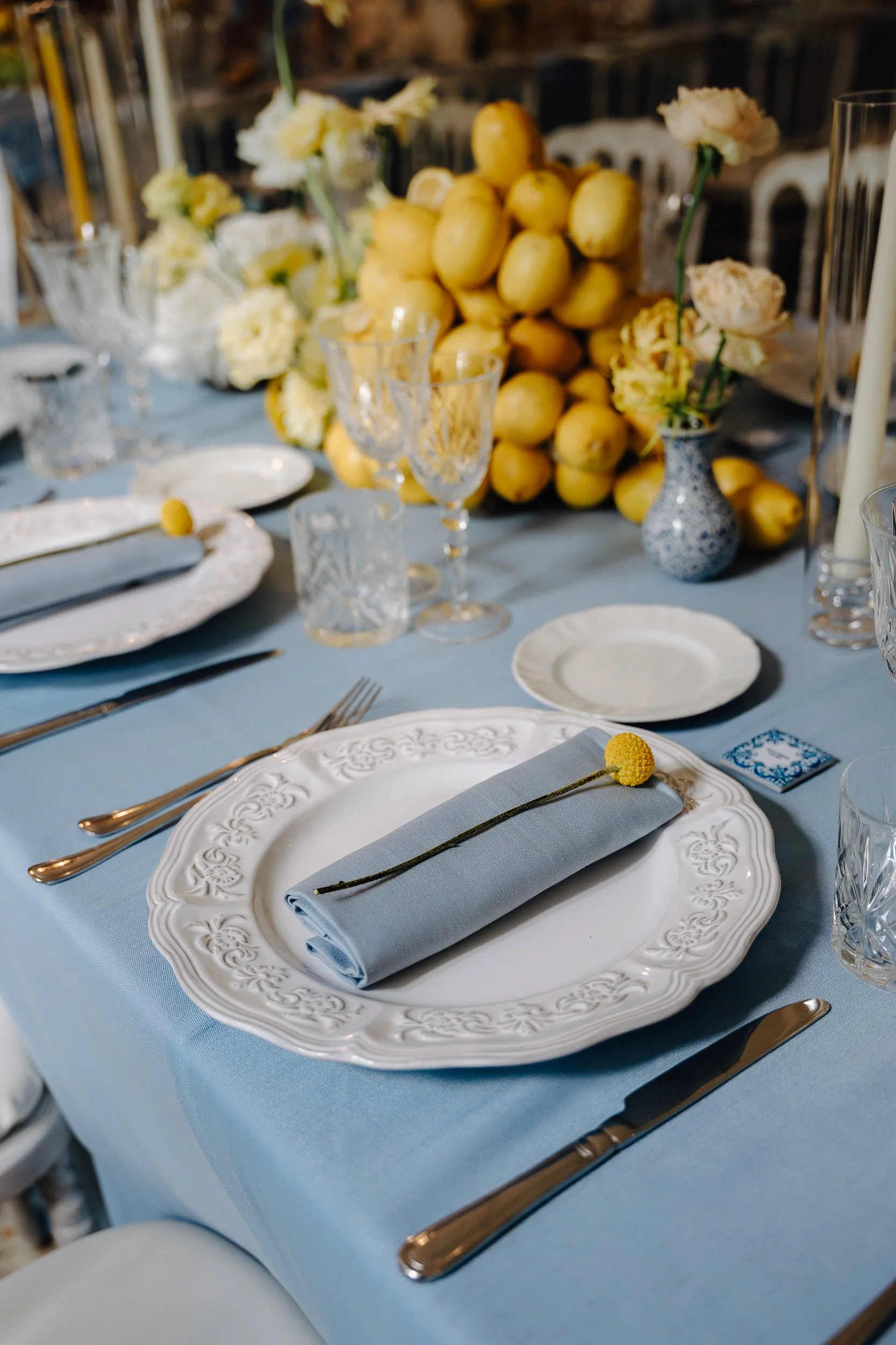 A formally set dining table with white ornate plates, a light blue napkin with a yellow flower on top, crystal glassware, and gold utensils, with a centerpiece of yellow fruits and white flowers in vases.