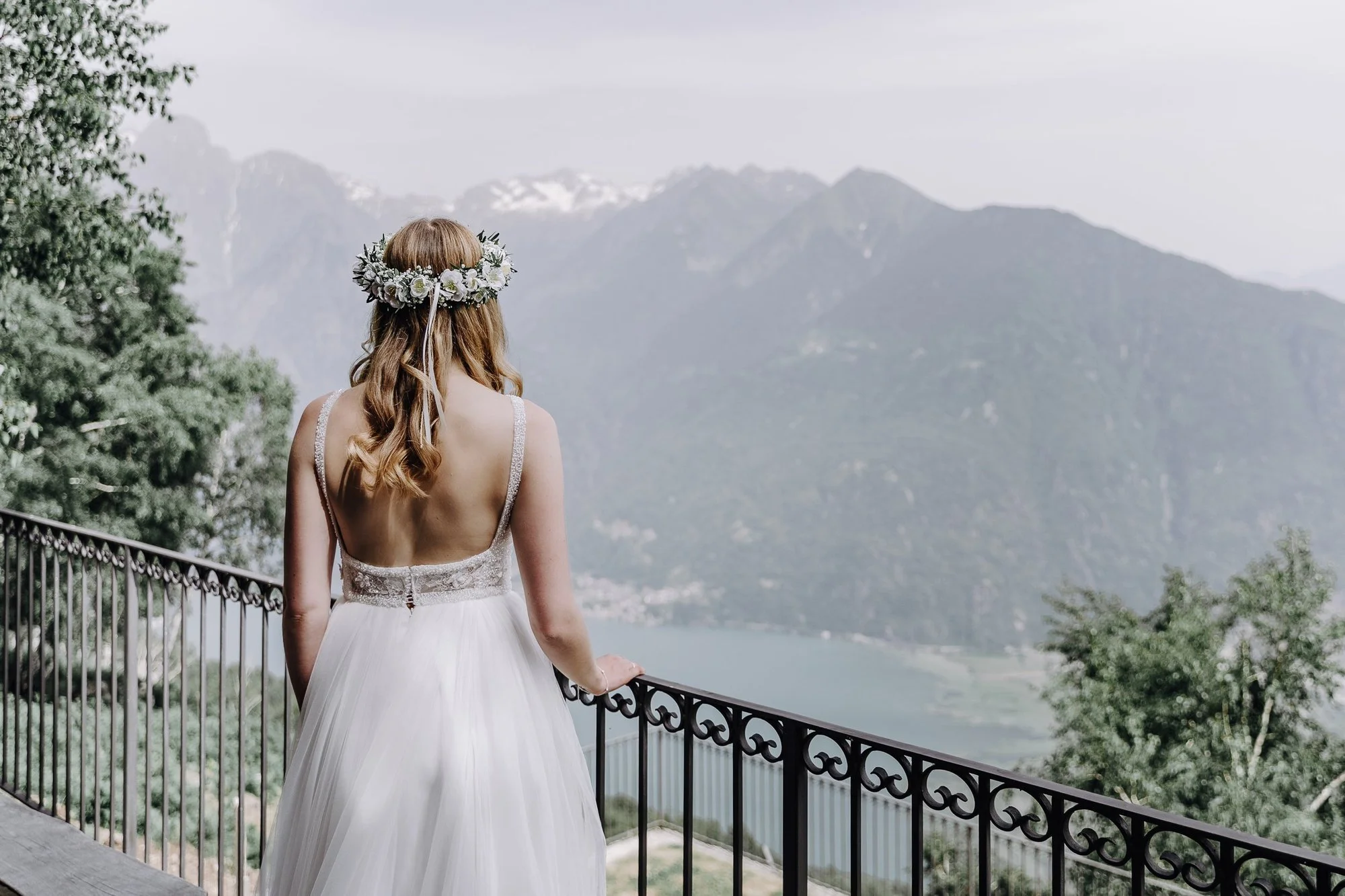 A woman in a white dress with a floral crown looks out over a mountainous landscape from a balcony.