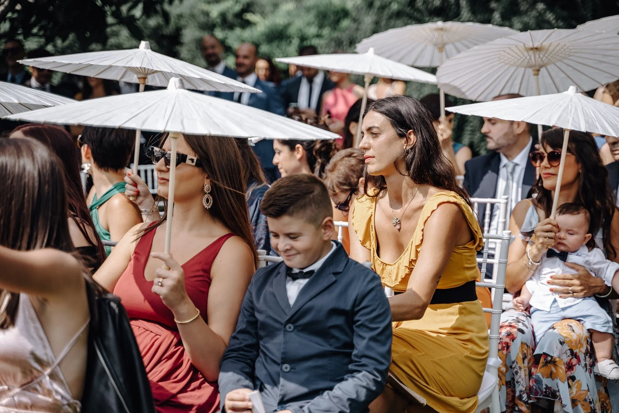 Group of people attending an outdoor event, sitting on white chairs under white umbrellas, with some in formal attire, among lush green trees.