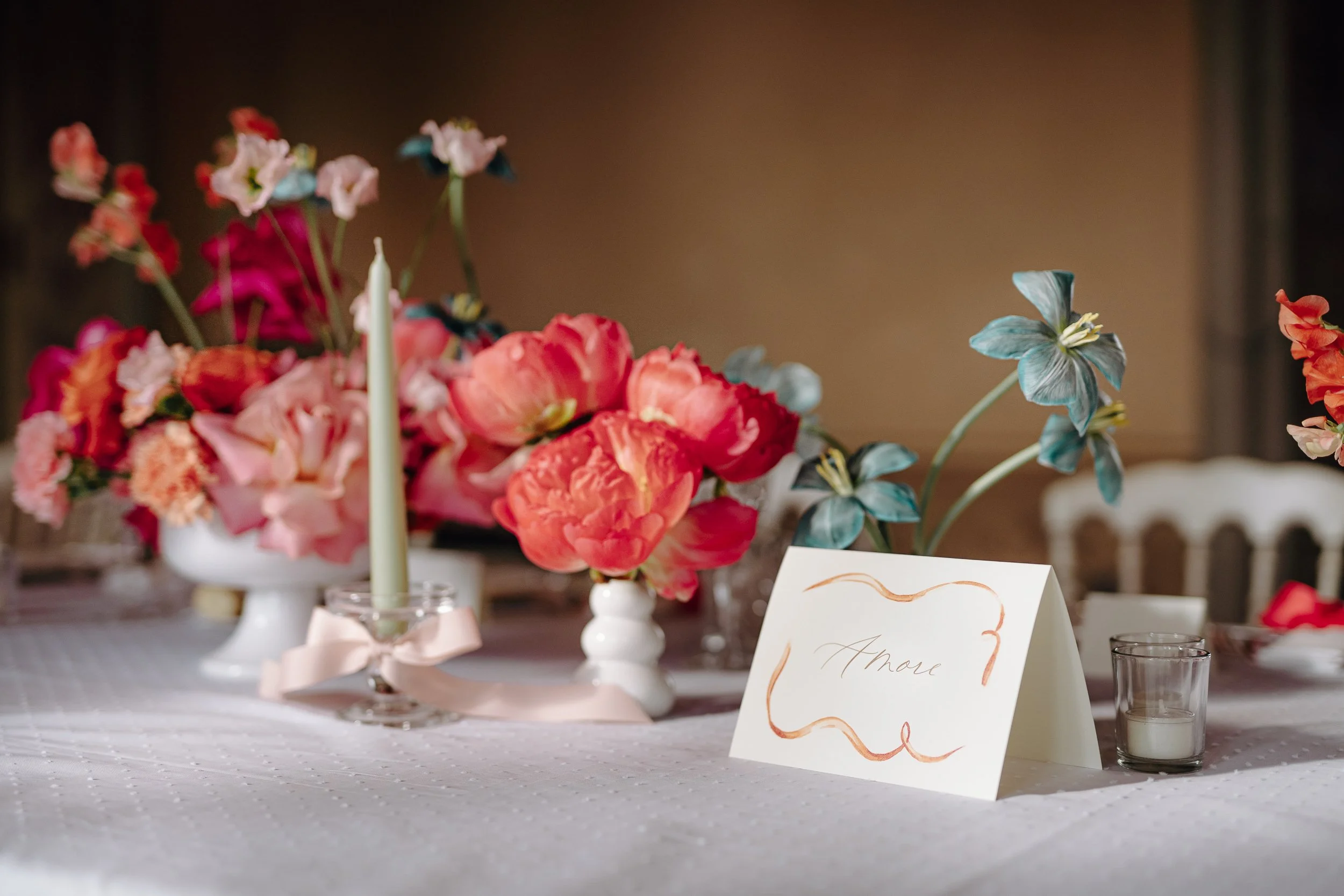 A floral centerpiece with pink, red, and purple flowers in white vases on a table, with a handwritten place card in the foreground and candles nearby.