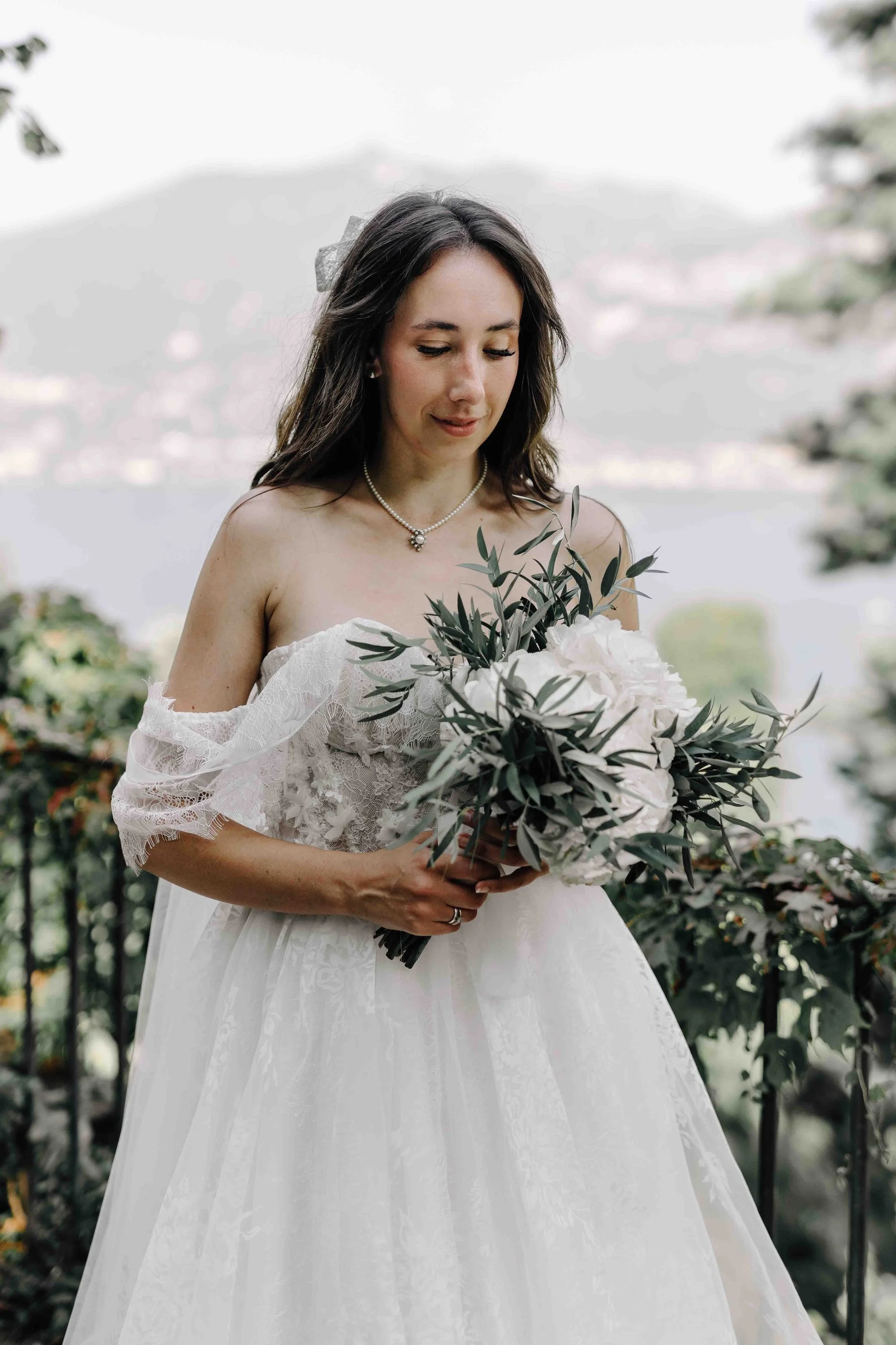 A woman in a white wedding dress holding a bouquet of white flowers and green leaves, standing outdoors with trees in the background.