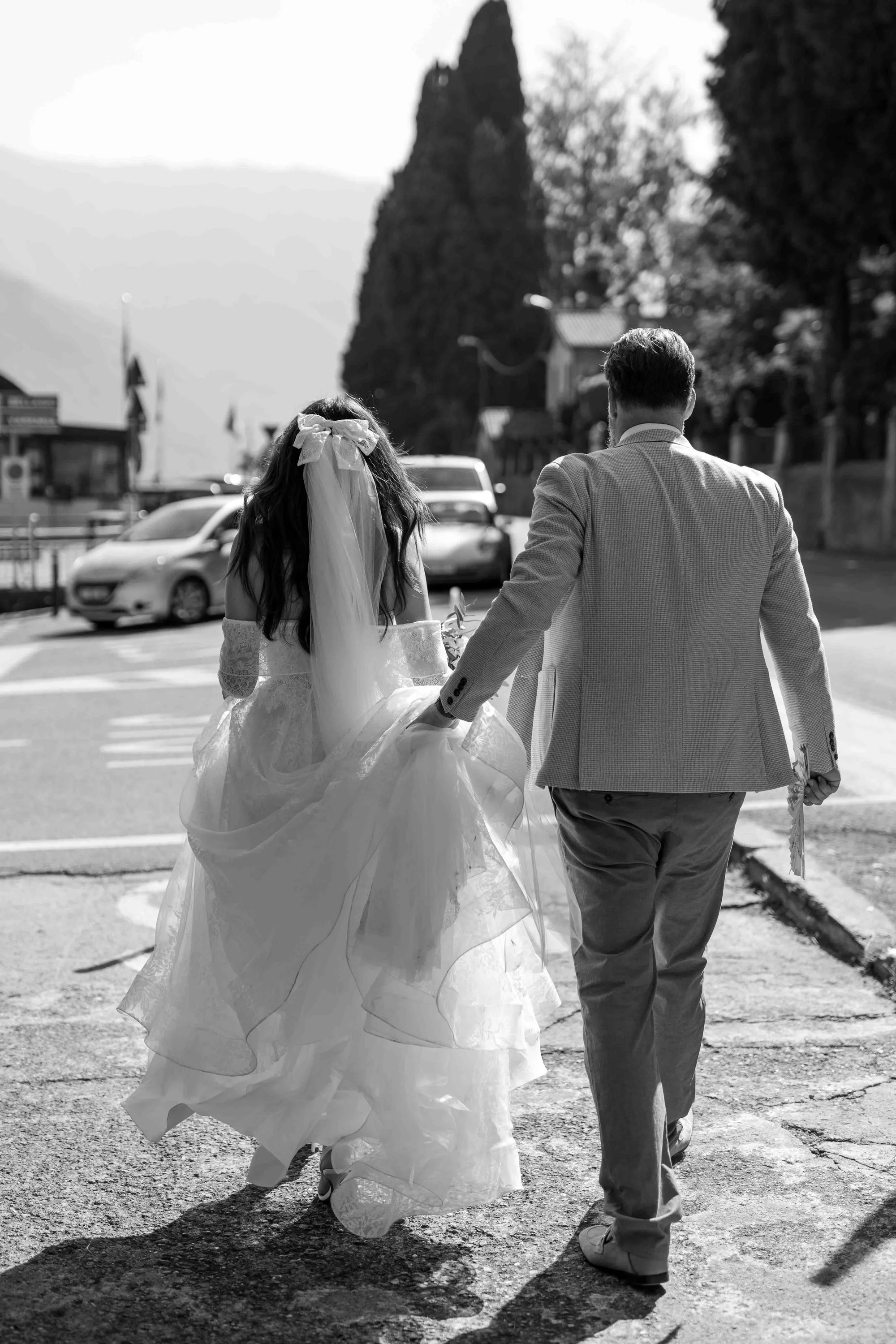 A man and a woman, dressed in wedding attire, are walking hand in hand outdoors on a sunny day. The woman wears a traditional wedding gown with a veil and bow, while the man is dressed in a light-colored suit. They are walking on a street with cars p