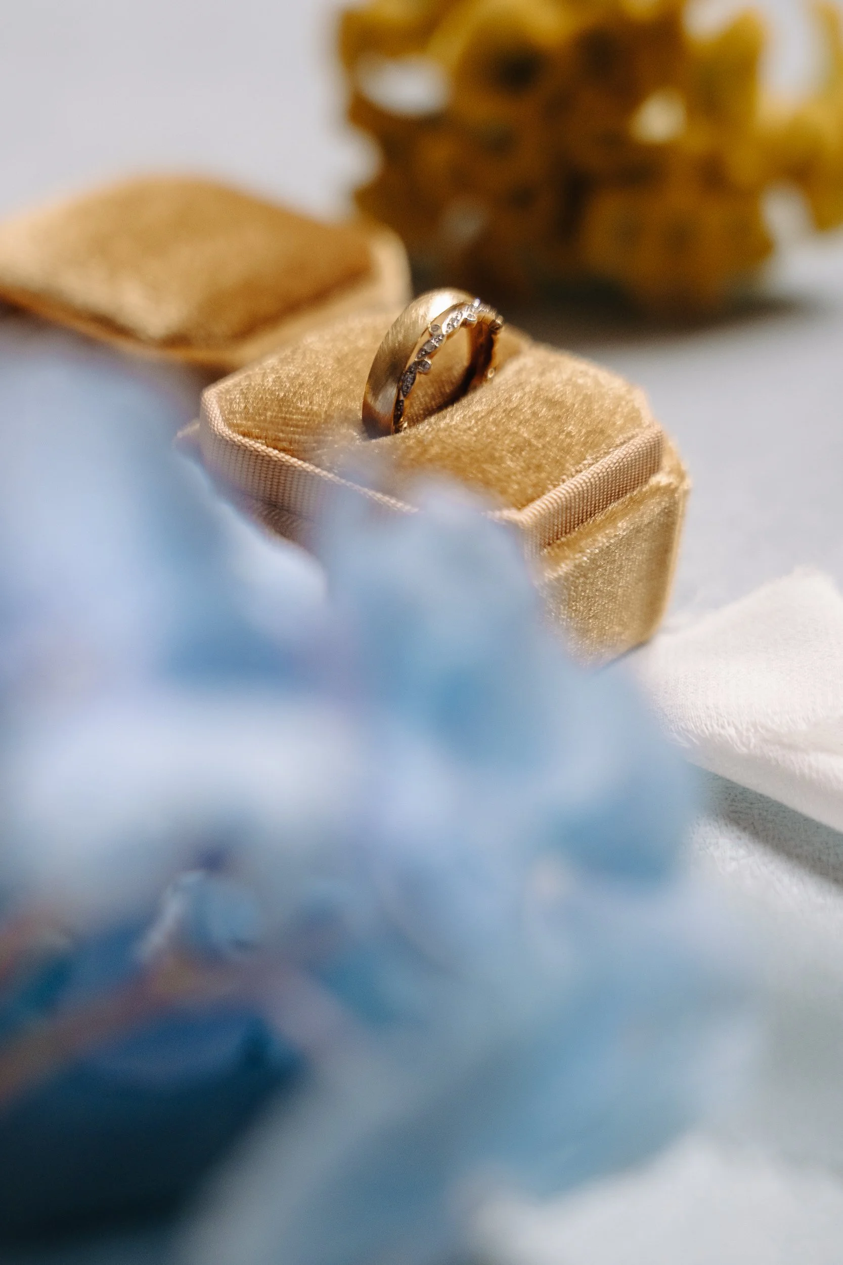 A gold wedding band with diamonds rests on a cream velvet ring box, with a blurred gold flower in the background.