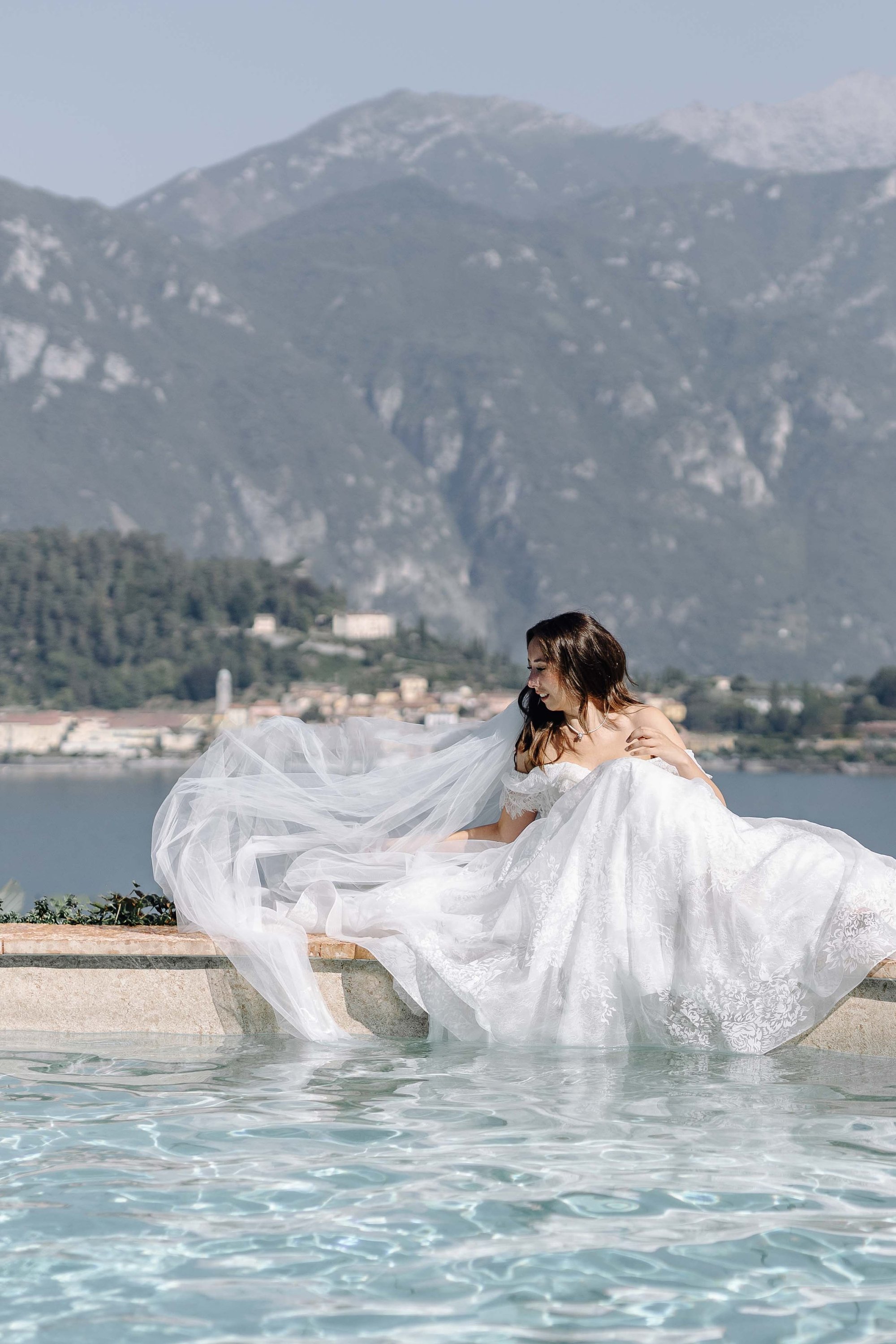 A woman in a wedding dress sitting on a ledge by a body of water with mountain scenery in the background.