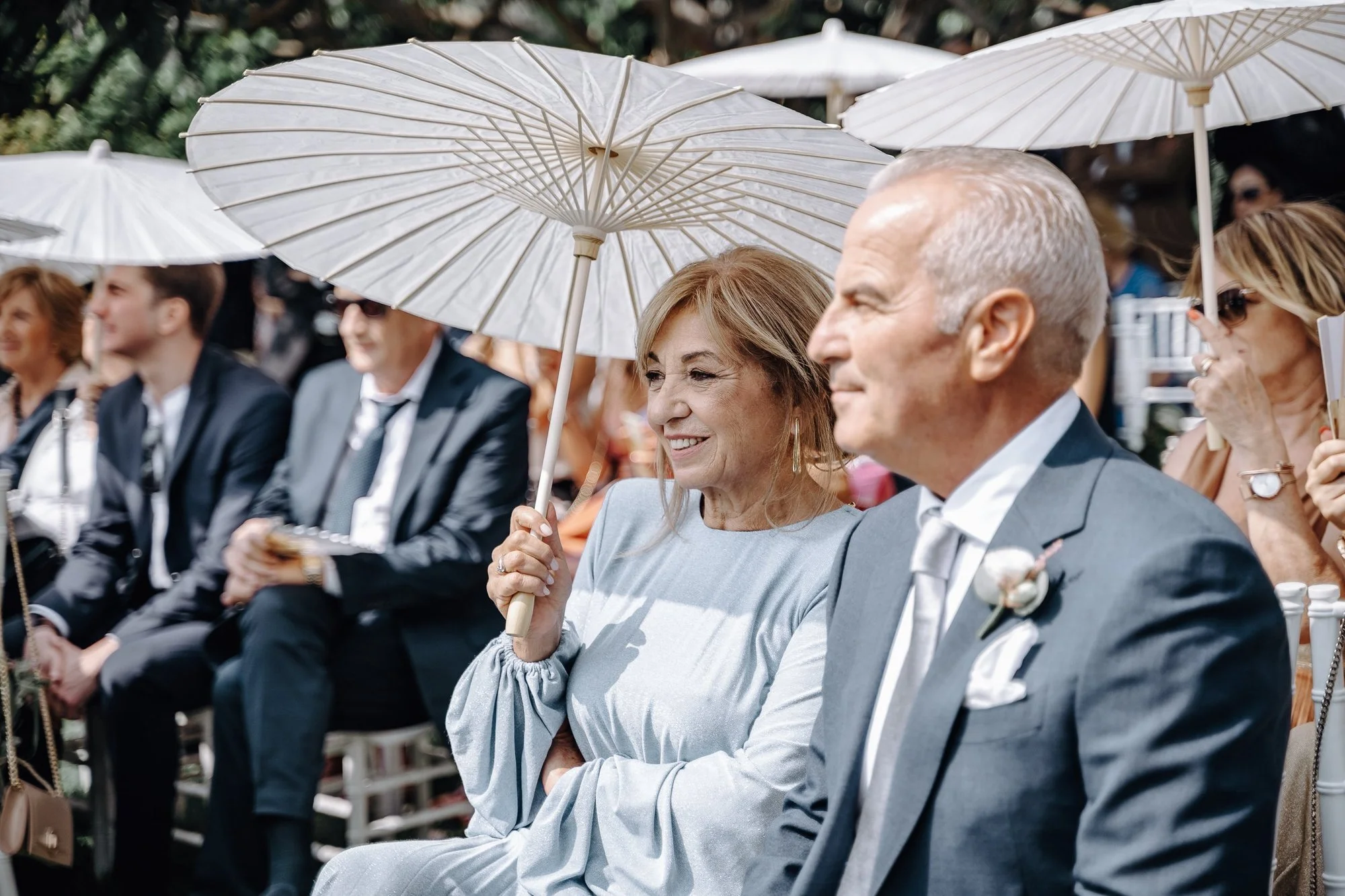 People seated outdoors at an event, holding white paper parasols. The focus is on a smiling middle-aged woman with light brown hair holding a parasol, and a man with gray hair seated next to her. Other attendees are visible in the background.